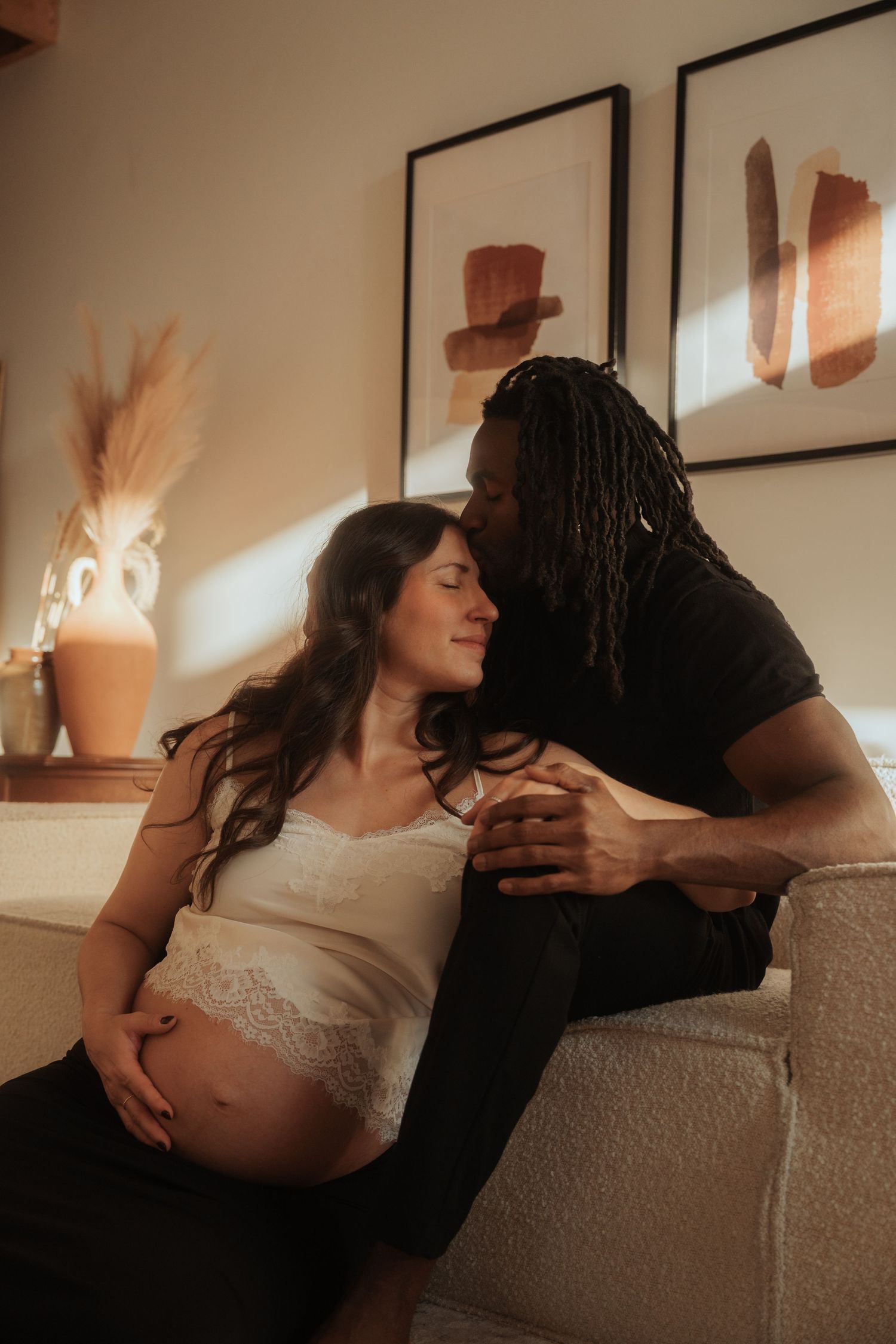 Couple sharing tender moment on couch with pregnant belly in warm light.