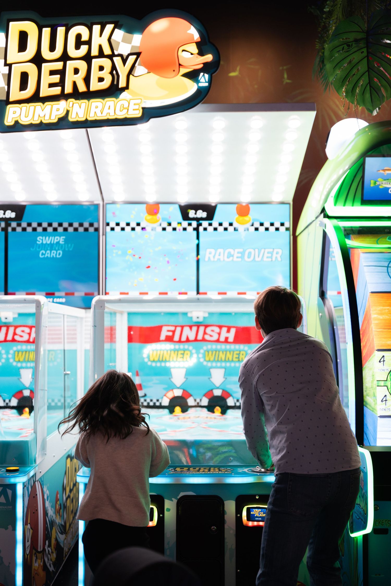Two children play Duck Derby Pump N Race arcade game at a colorful indoor arcade.