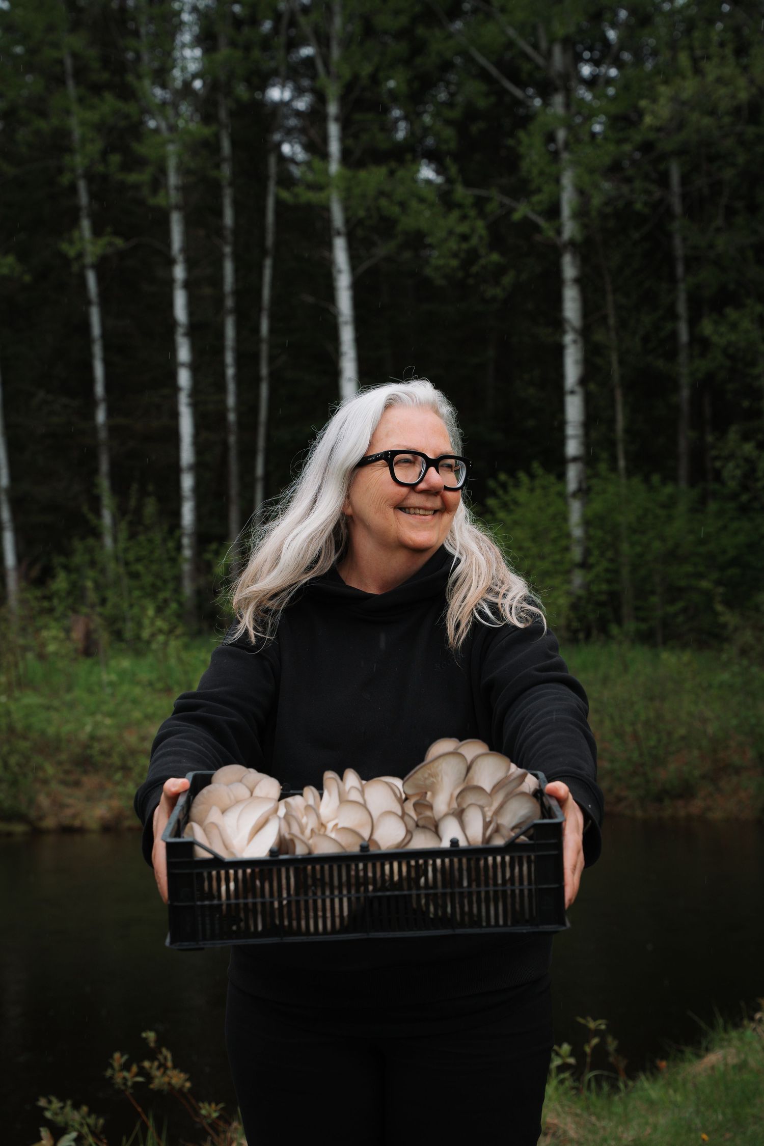 Smiling woman with white hair holding a crate of fresh oyster mushrooms outdoors near trees.