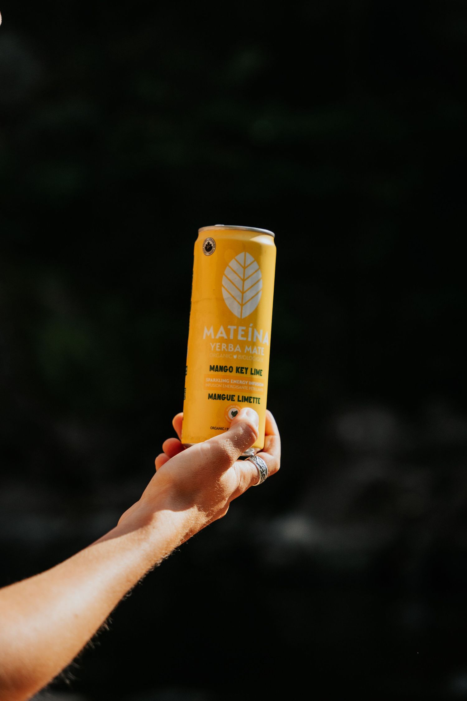 Hand holding yellow Matein Mango Key Lime sparkling drink can against dark background.
