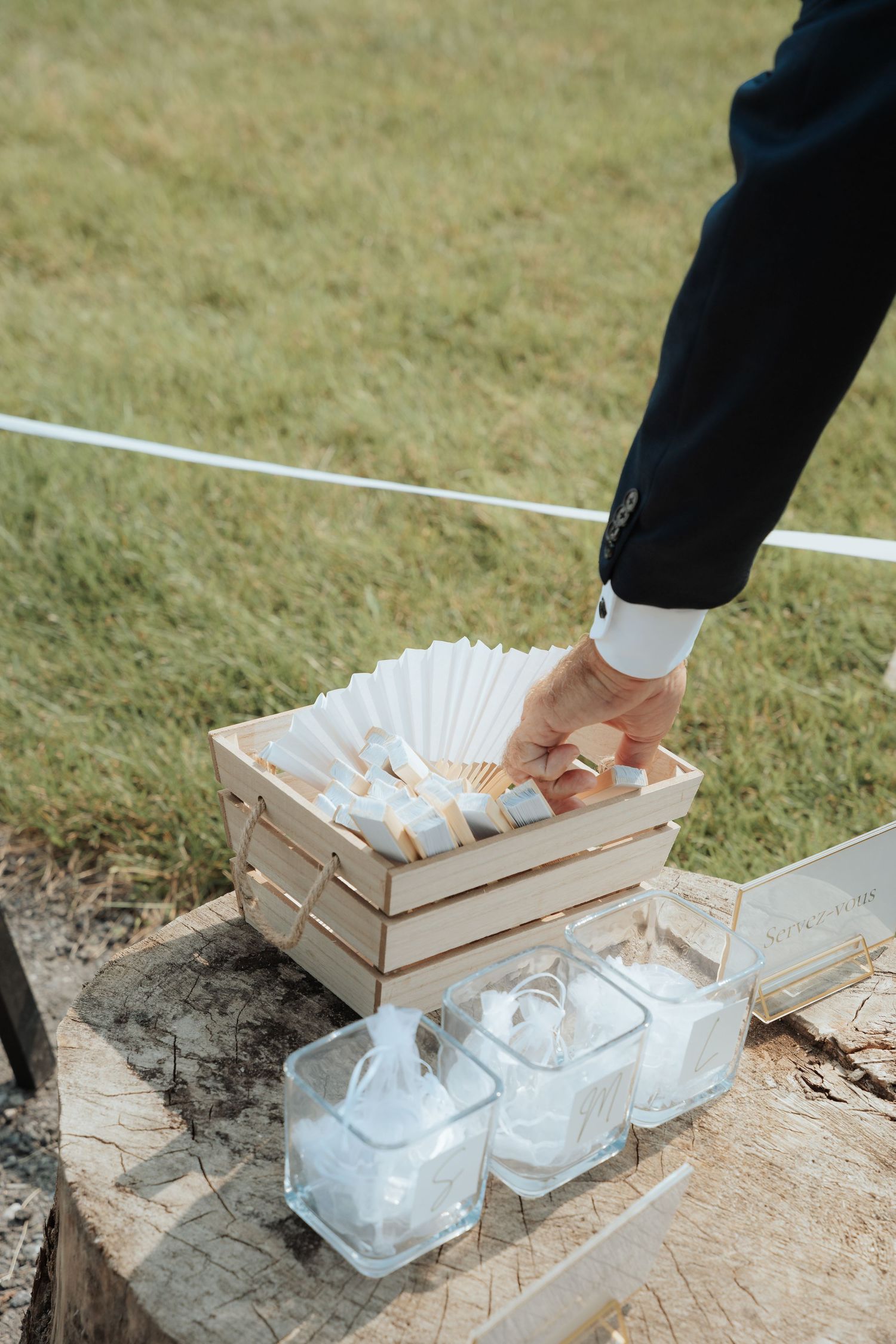 A groom in a navy suit reaches into a wooden crate filled with white paper fans on a tree stump at an outdoor wedding.