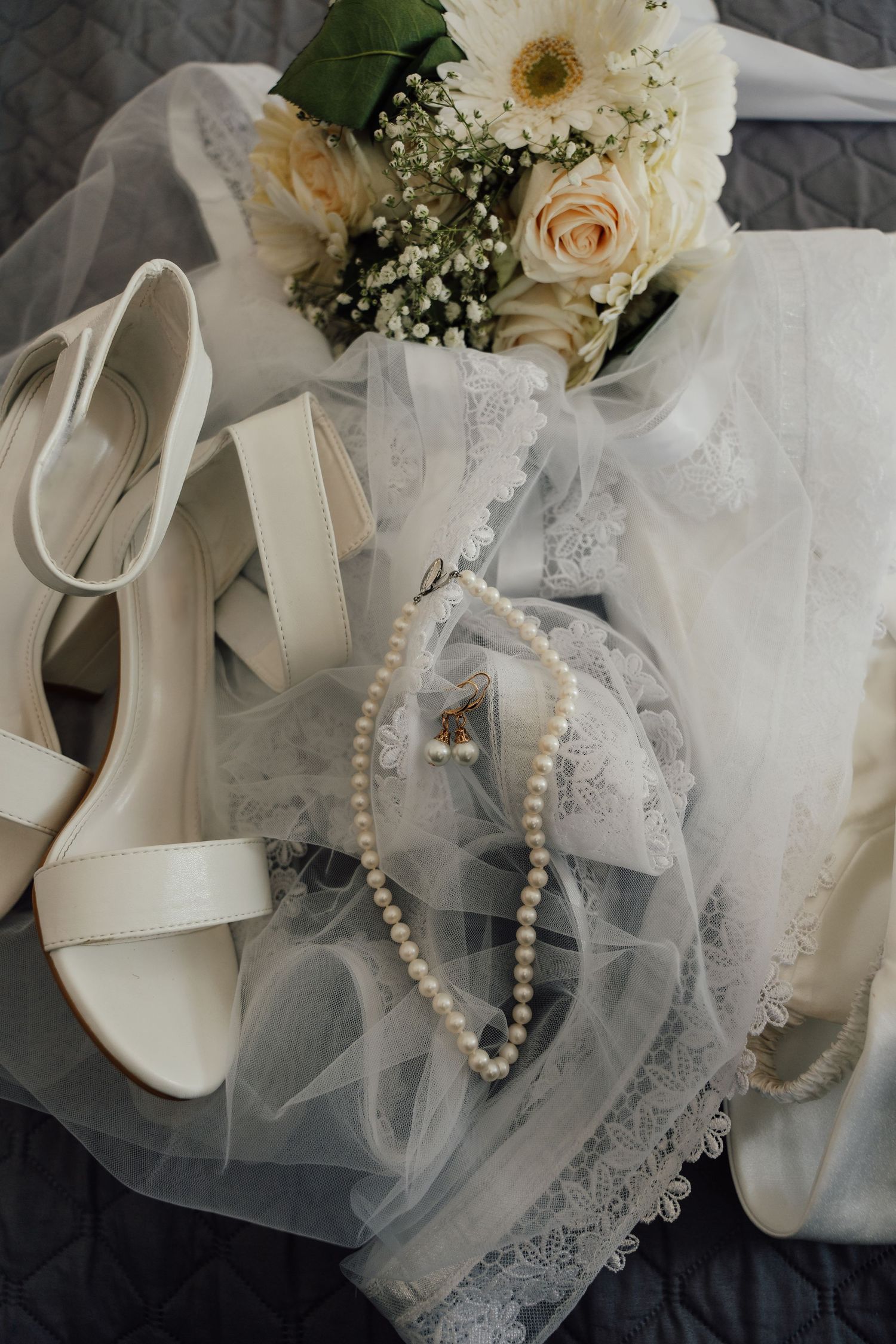 Bridal flat lay featuring white heels, pearl necklace, lace veil, and a bouquet of roses and wildflowers.