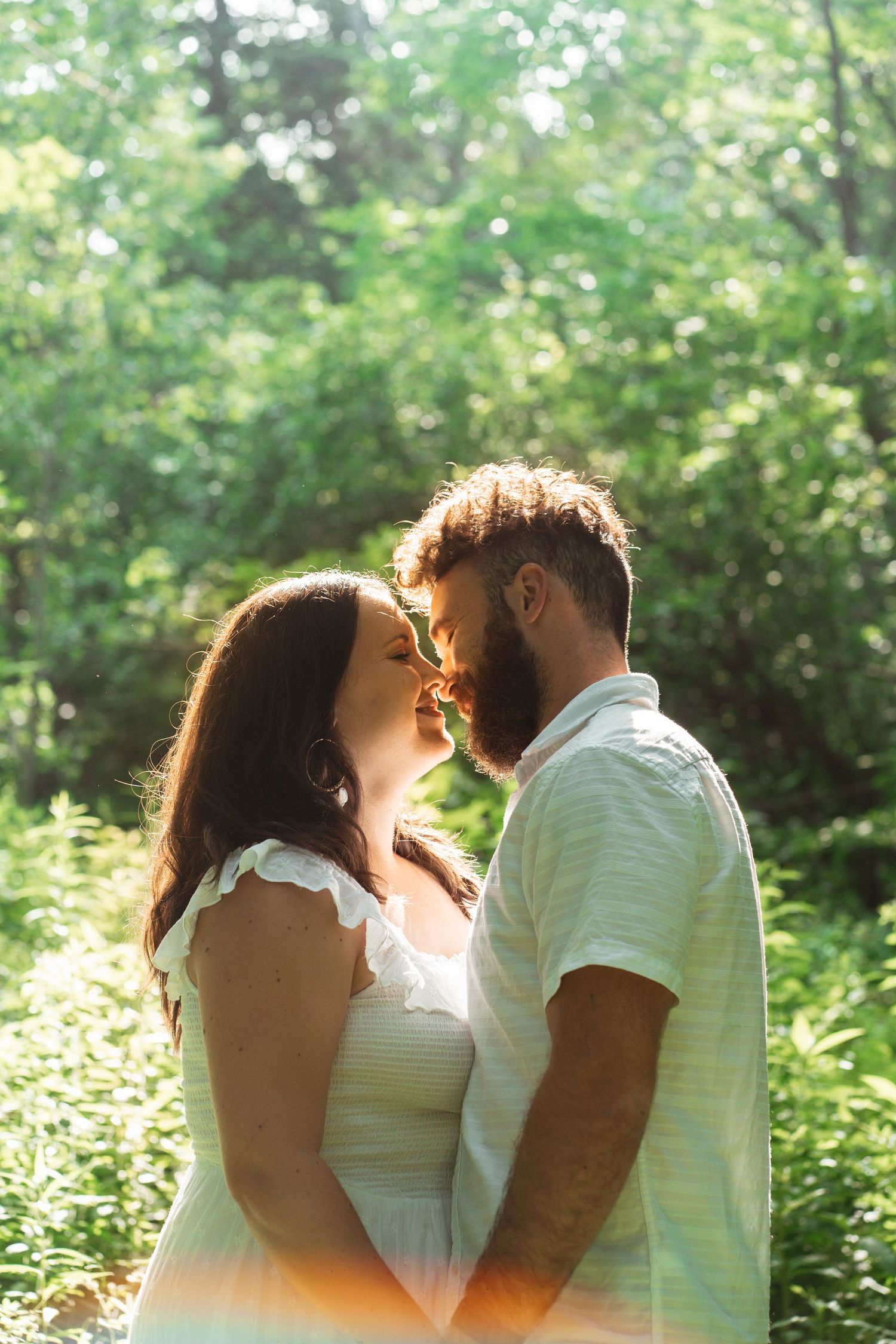 Couple sharing a romantic almost-kiss moment surrounded by lush green forest in warm sunlight.