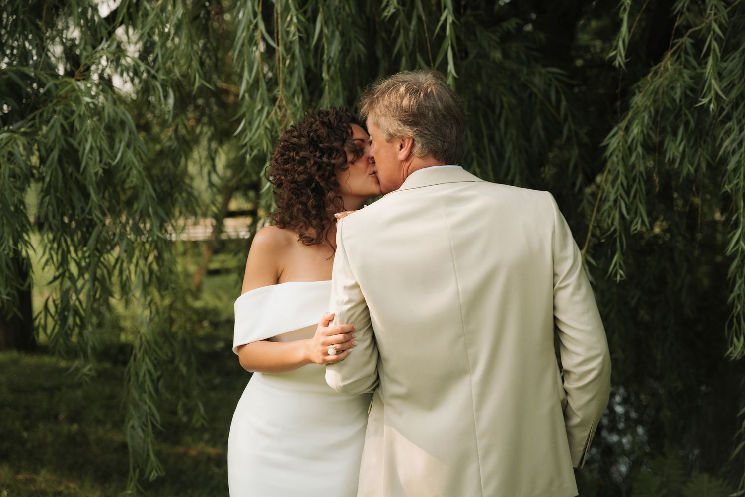 Newlywed couple shares a romantic kiss under weeping willow branches in a garden setting.