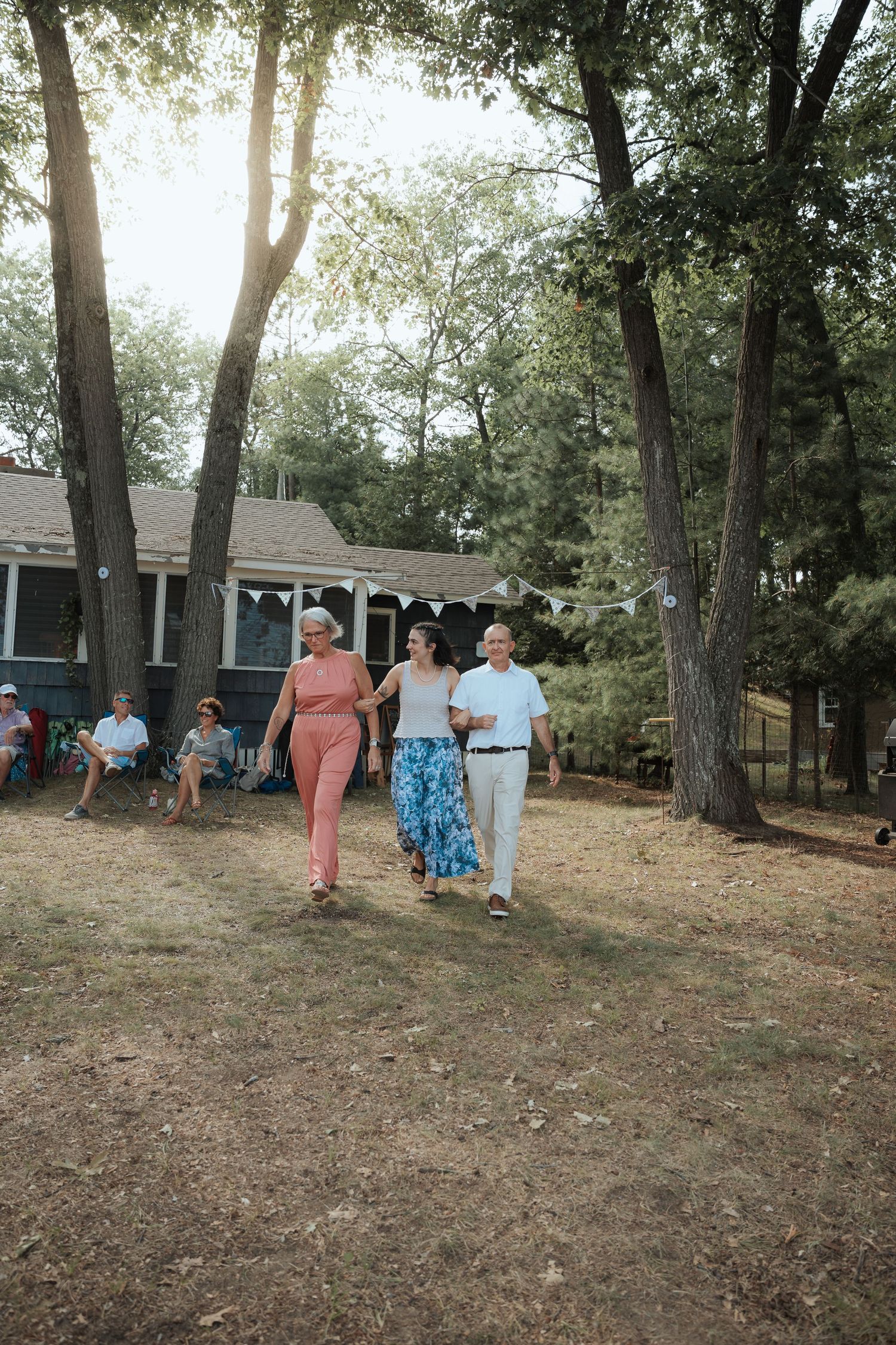 Three guests walk together through a sunlit backyard at an outdoor wedding reception surrounded by tall trees.
