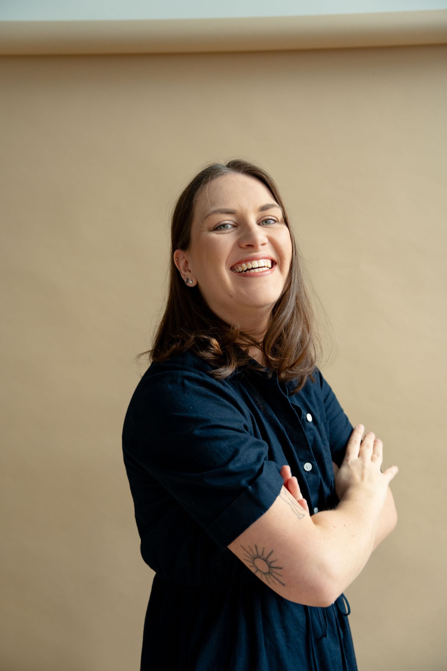 Smiling brunette woman with tattoo posing with arms crossed in navy dress.