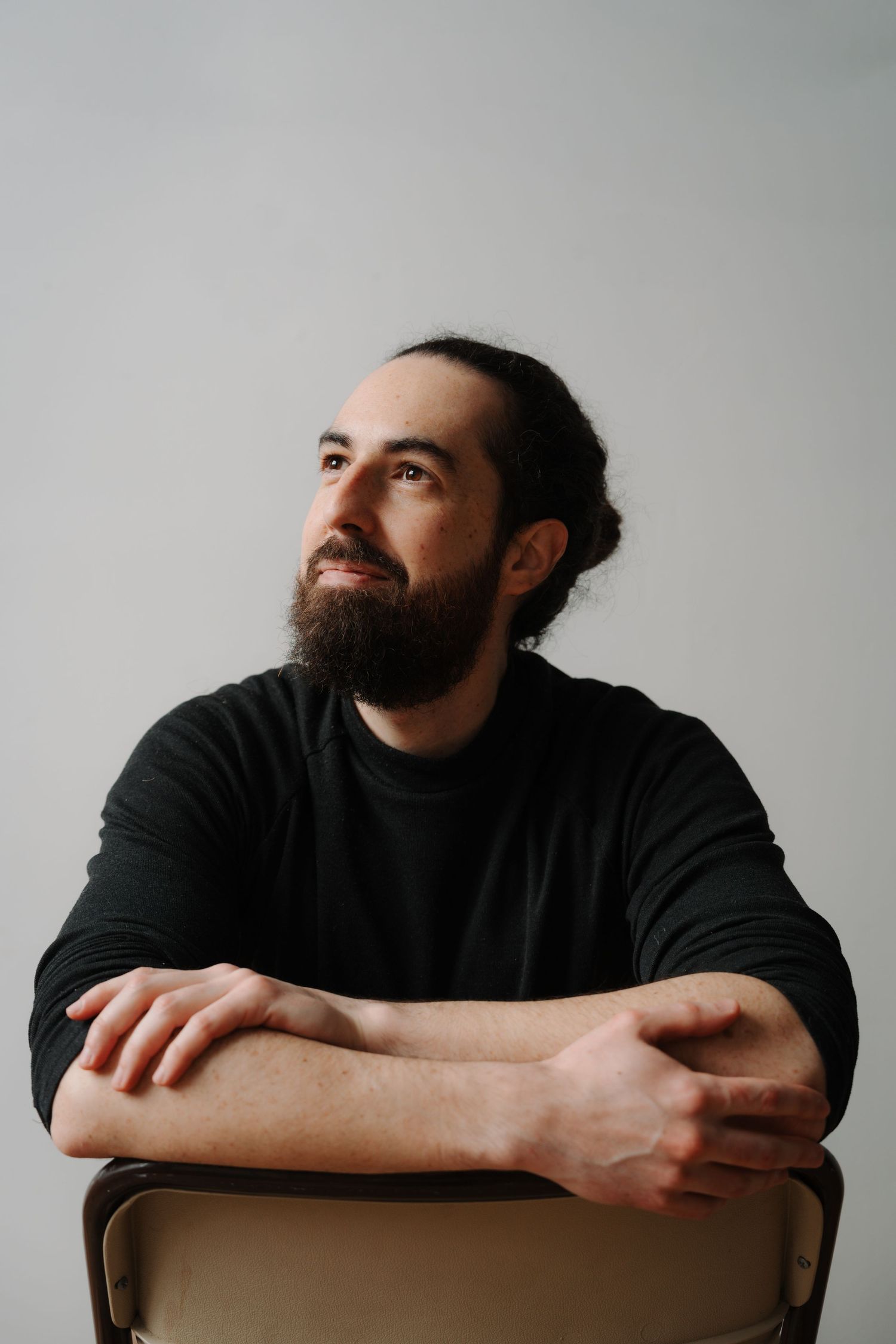 Bearded man with long dark hair sitting on chair with arms crossed, looking up thoughtfully.