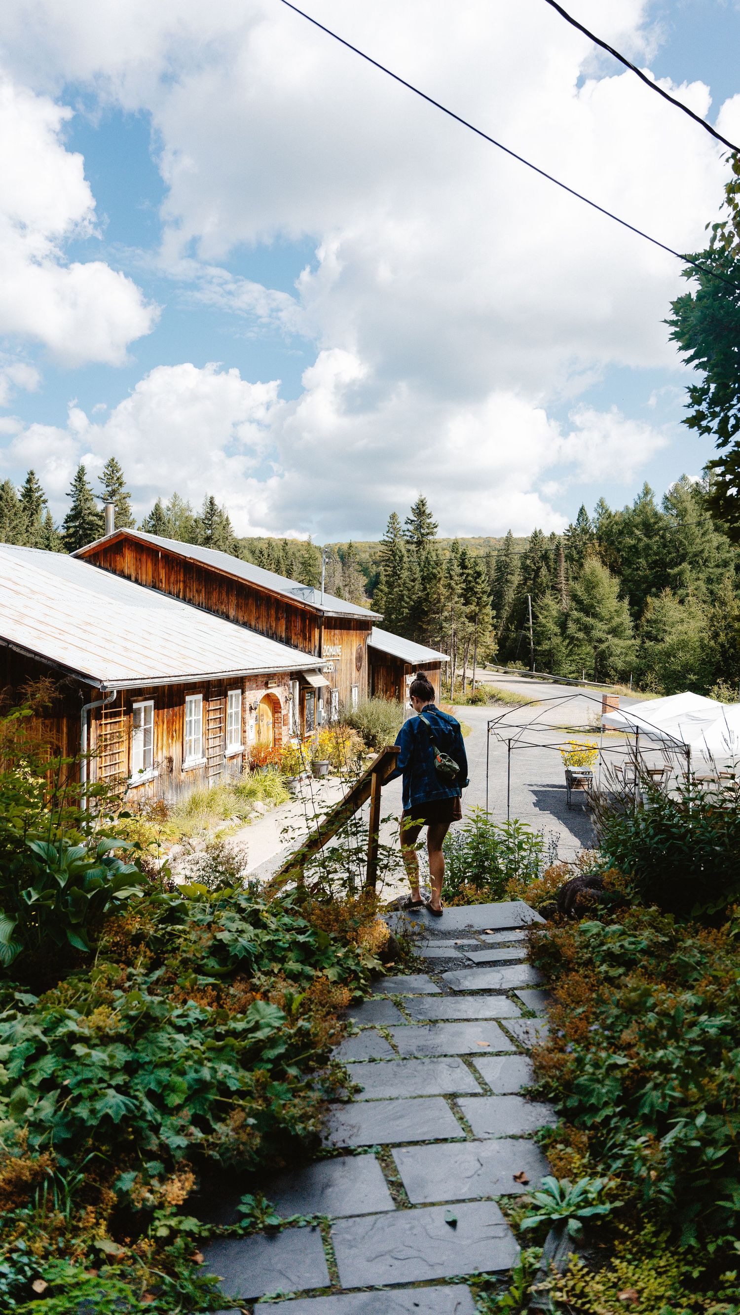 Person with backpack walking along a stone path beside a rustic wooden building in a forest.
