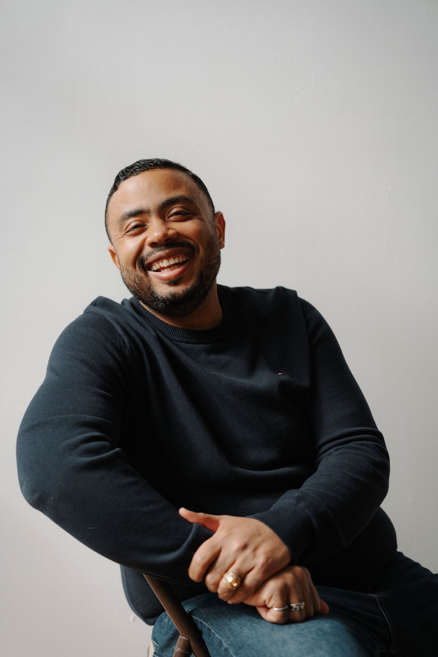 Happy man in black sweatshirt laughing while seated against a light gray background.