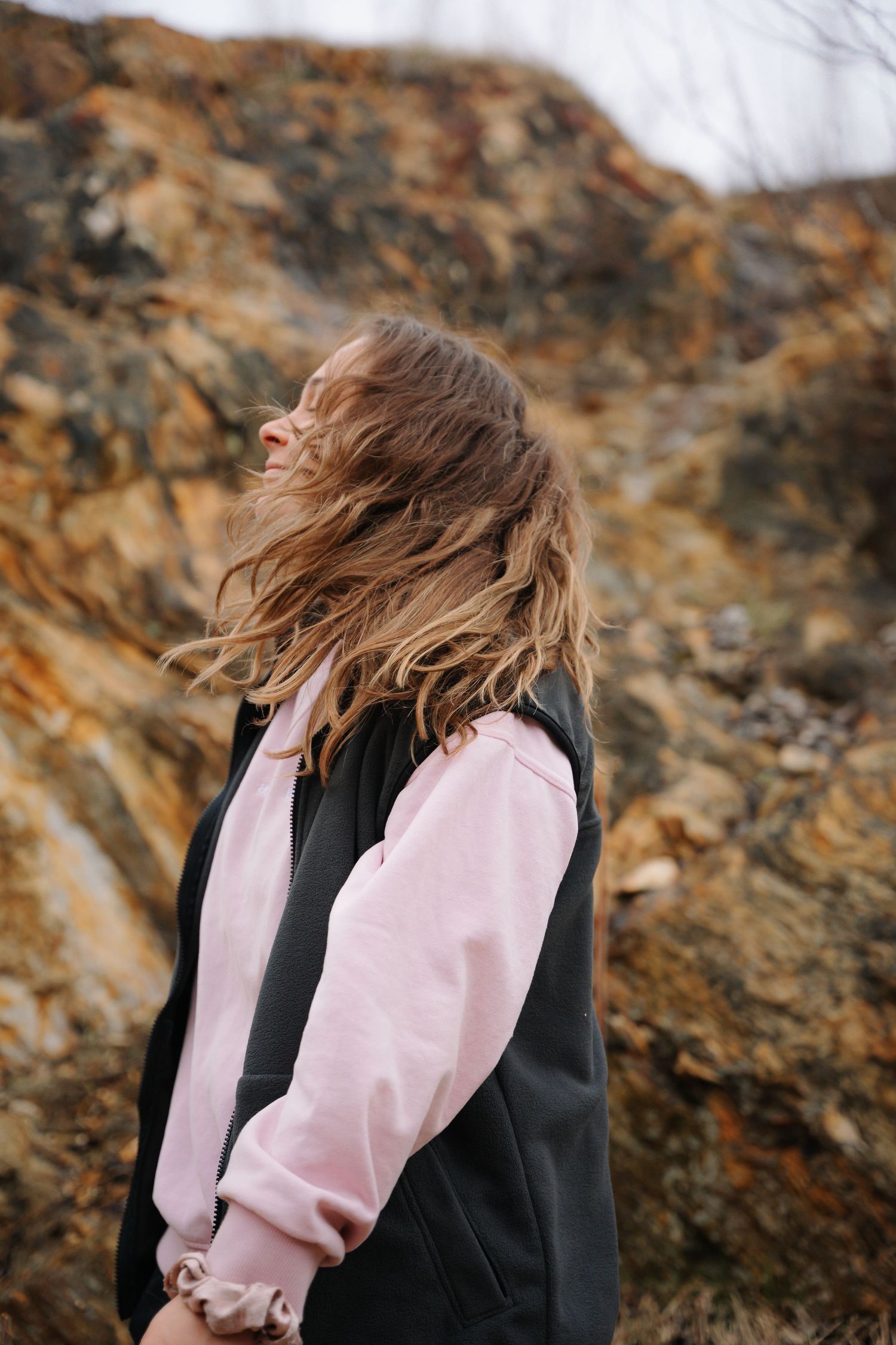 Young woman with windswept hair standing against rugged rocky cliff wearing pink and black jacket.