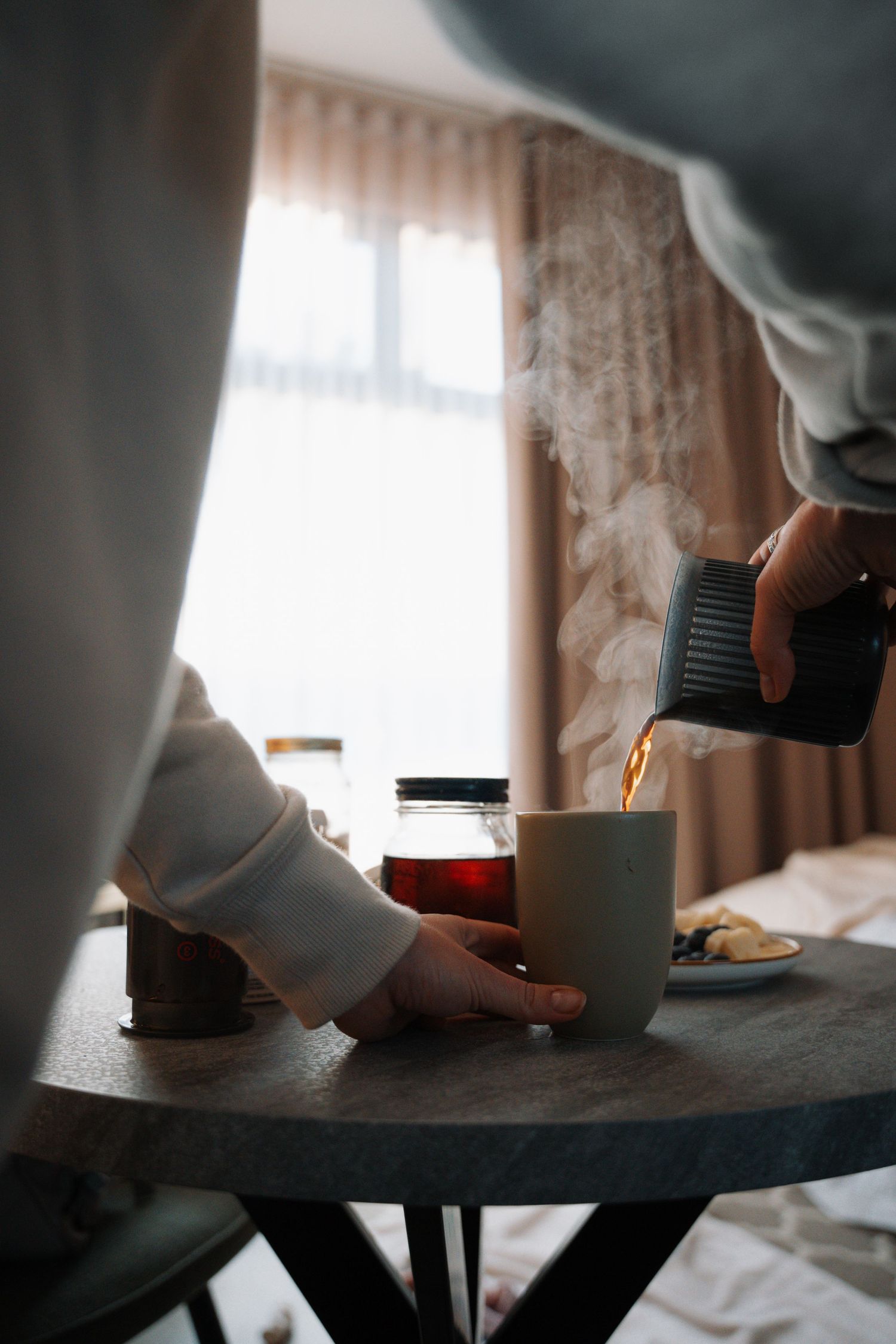 Person pouring hot steaming tea from a teapot into a white mug on a round wooden table.