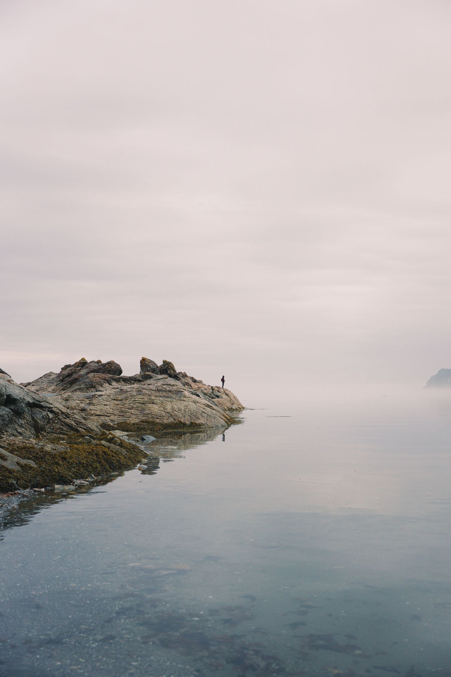 Lone figure standing on rocky shoreline in misty calm coastal landscape.