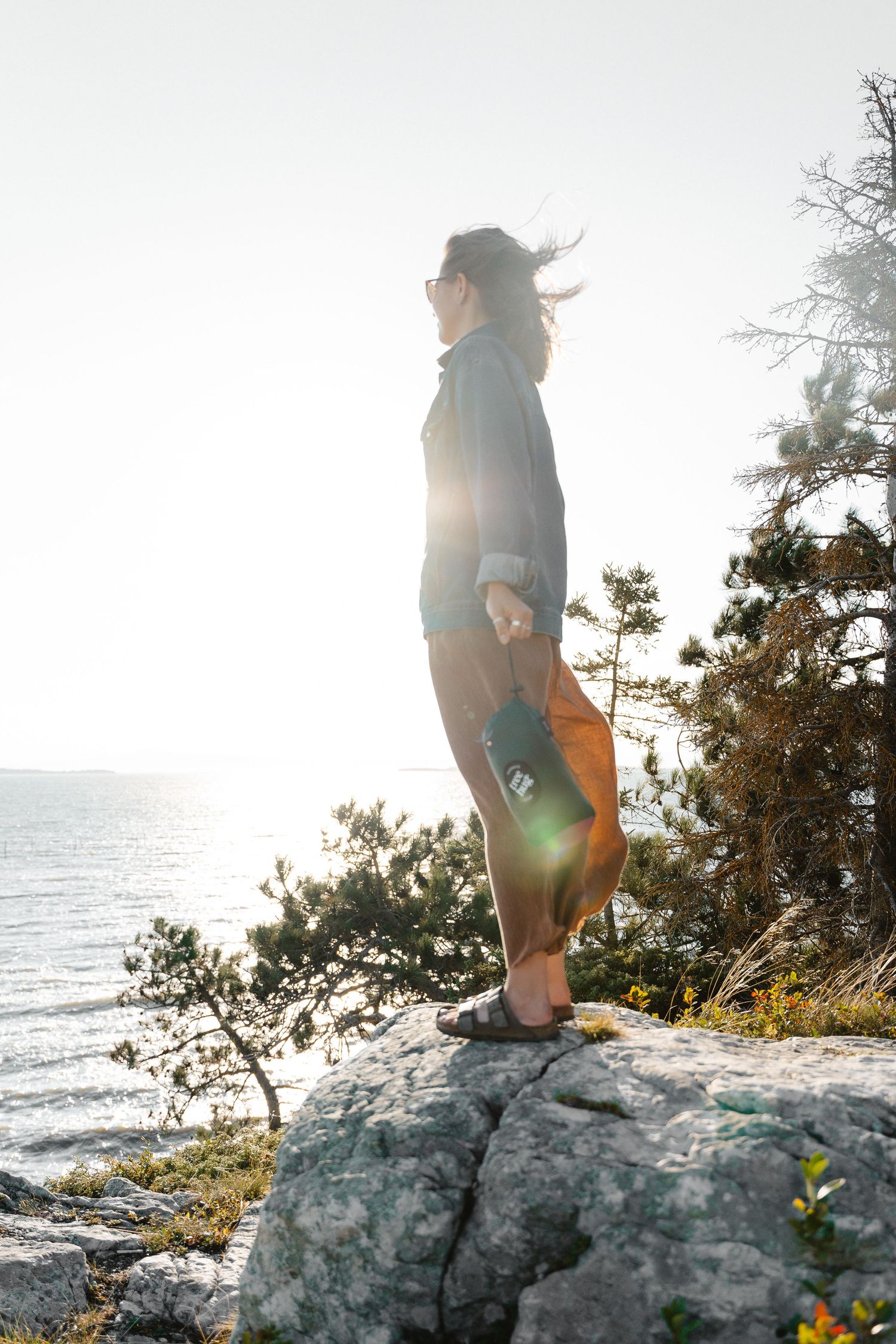 Woman standing on rocky cliff overlooking the ocean at sunset with wind blowing her hair.