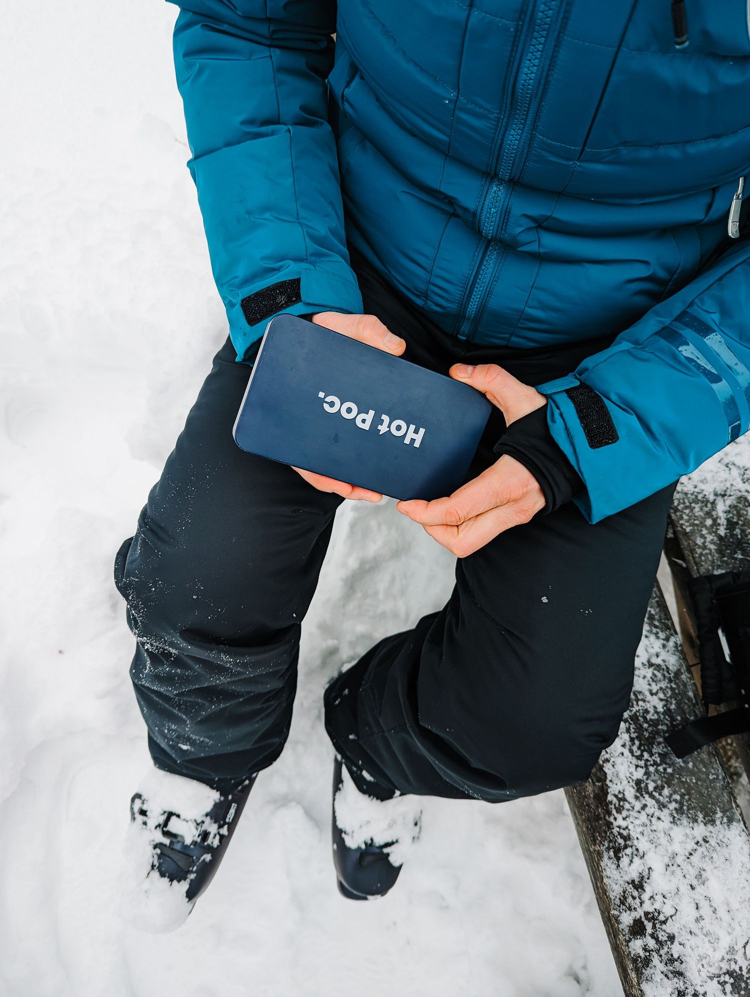 Person in blue ski jacket holding a navy Hot Poc hand warmer while sitting in snow.