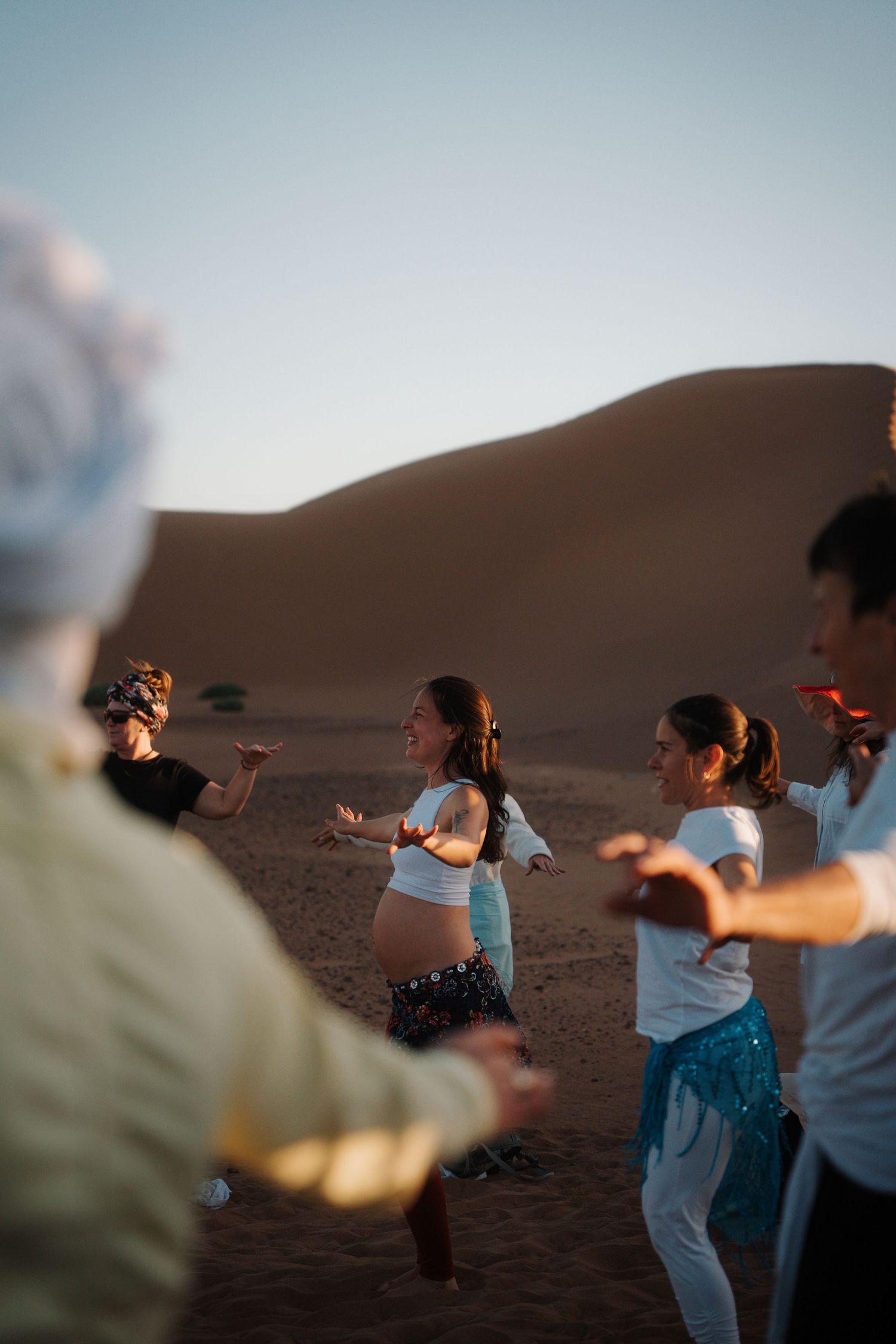 Group of people dancing in the Sahara desert at sunset with golden sand dunes in the background.