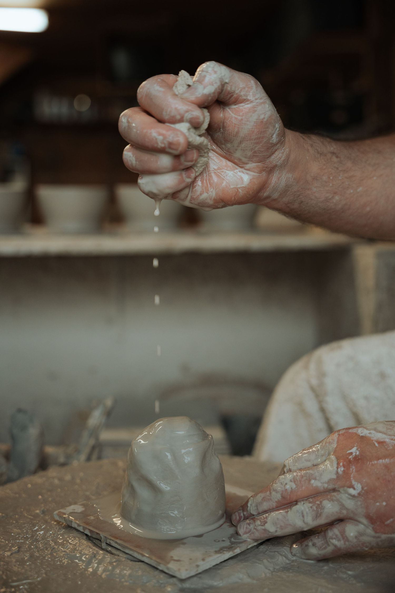 Close-up of clay-covered hands squeezing water over a small pottery piece on a work surface.