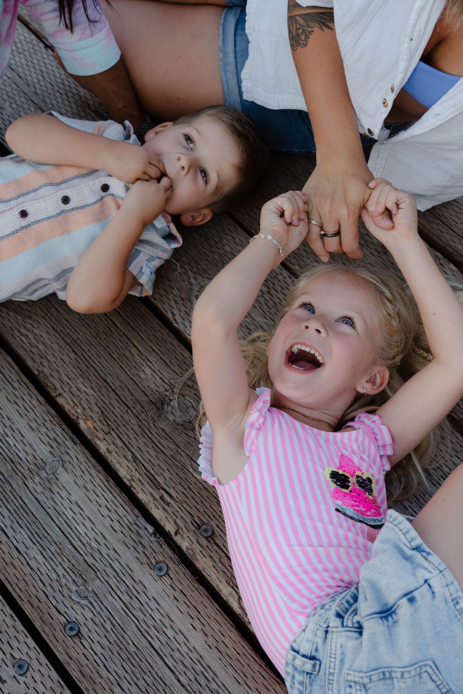 Two young girls laughing and lying on a wooden deck, holding hands with an adult woman standing above them.