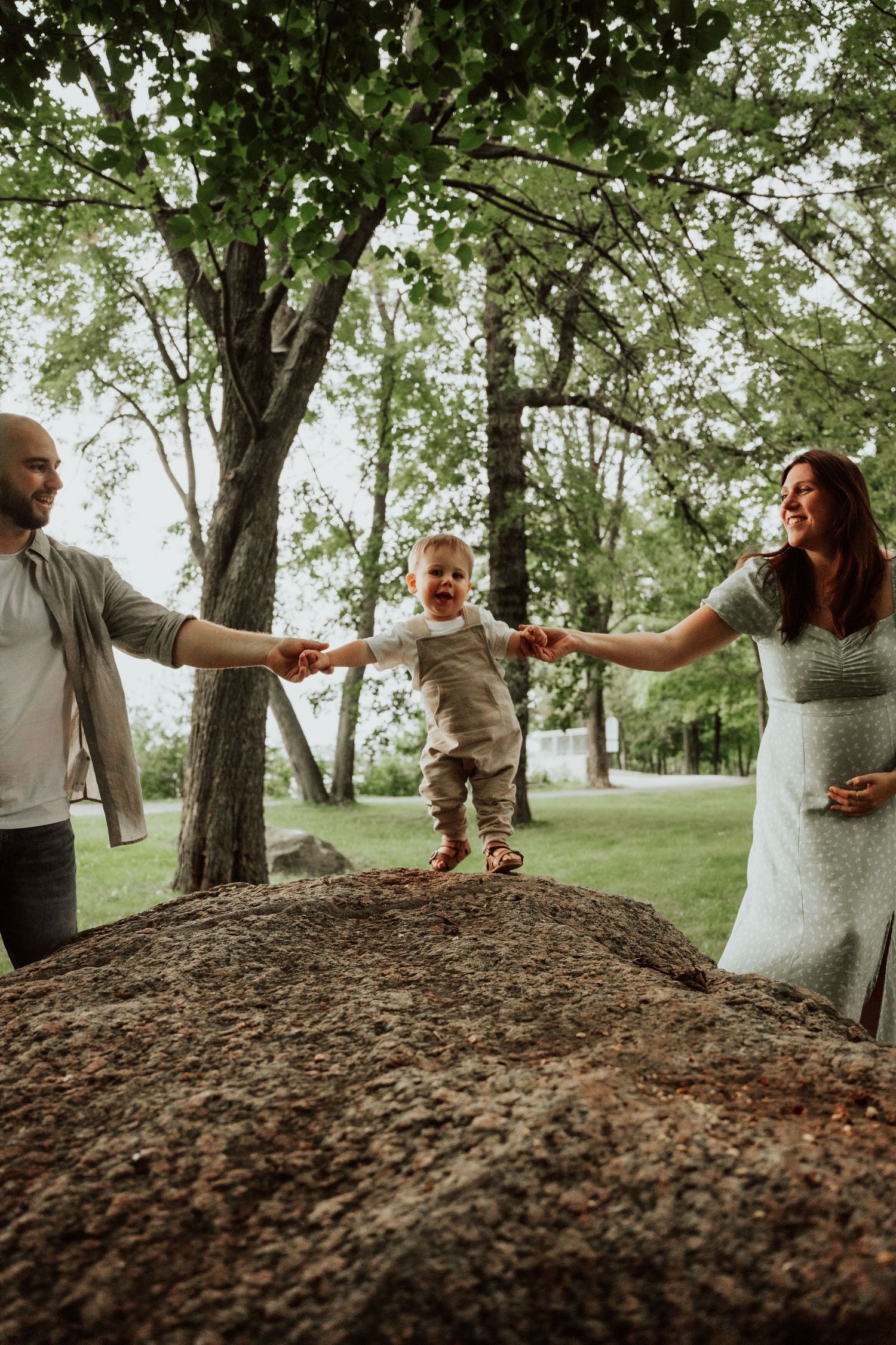 Parents hold a toddler's hands on a rock in a lush green park setting.