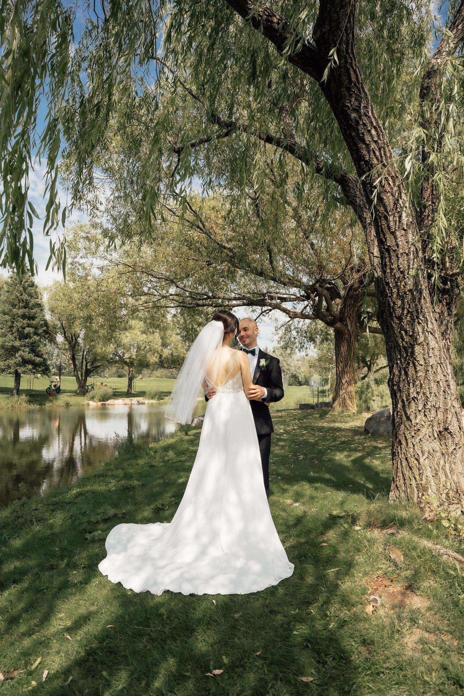 Bride in flowing white gown and veil stands with groom under a willow tree.