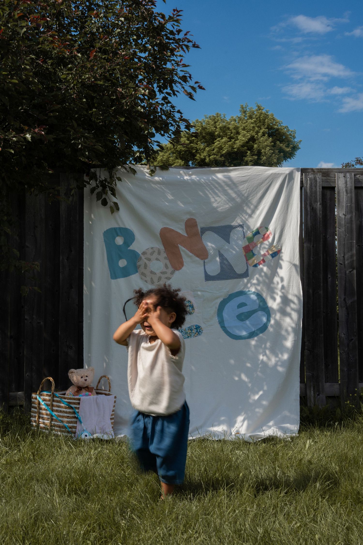 A happy toddler plays outdoors in front of a colorful handmade banner in a sunny backyard.