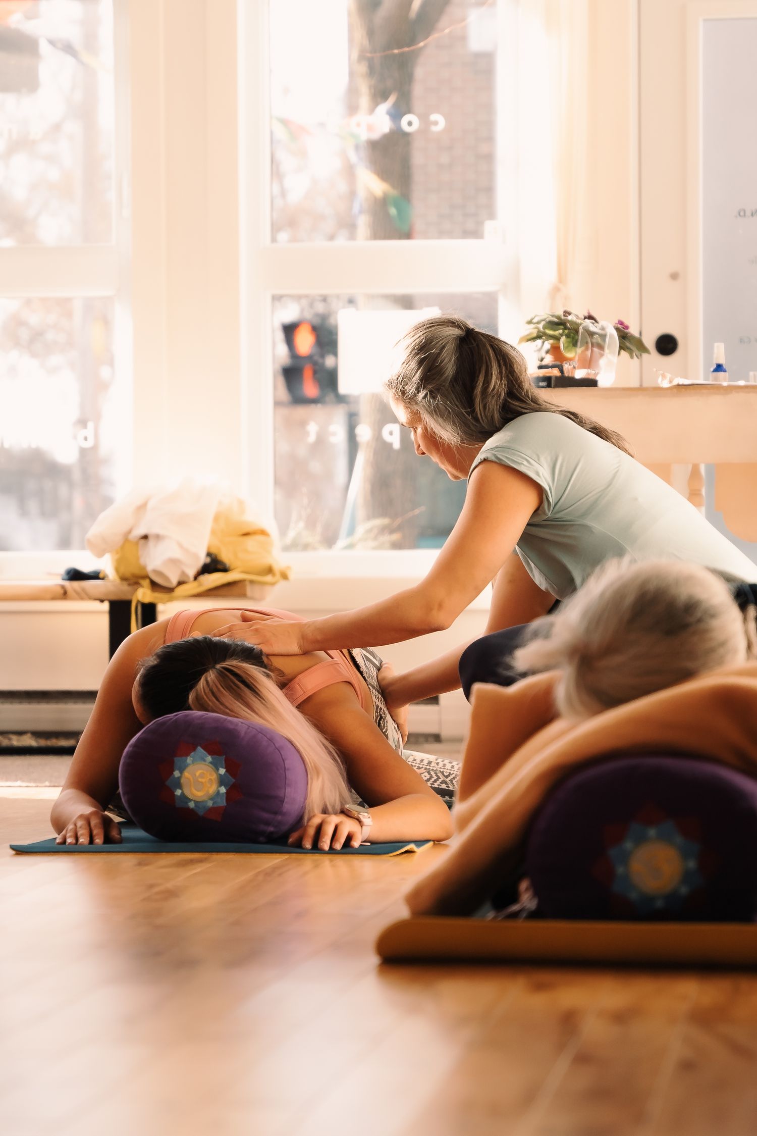 Yoga instructor assists a student in child's pose during a restorative yoga class on wooden floor.