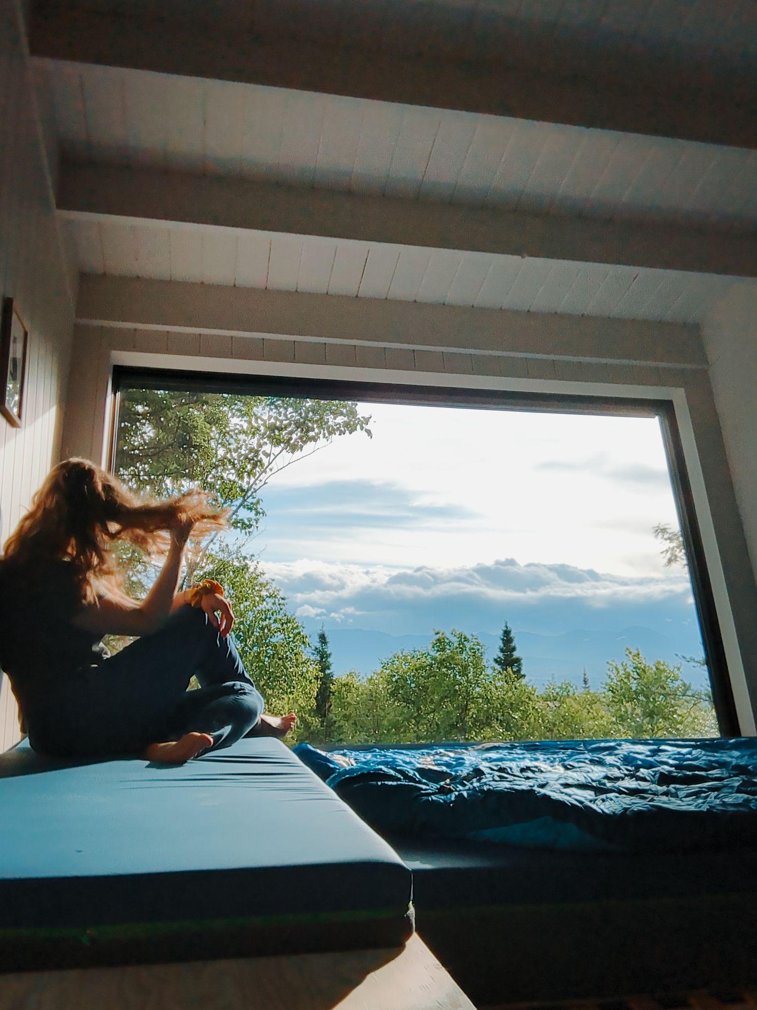 Woman sitting on bed by large panoramic window with scenic mountain and sky view in a cabin.