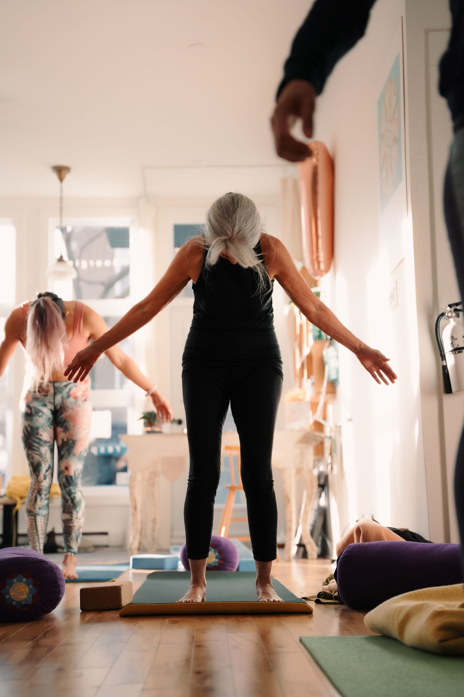 Senior woman in black standing on yoga mat with arms outstretched in a balance pose in a studio.