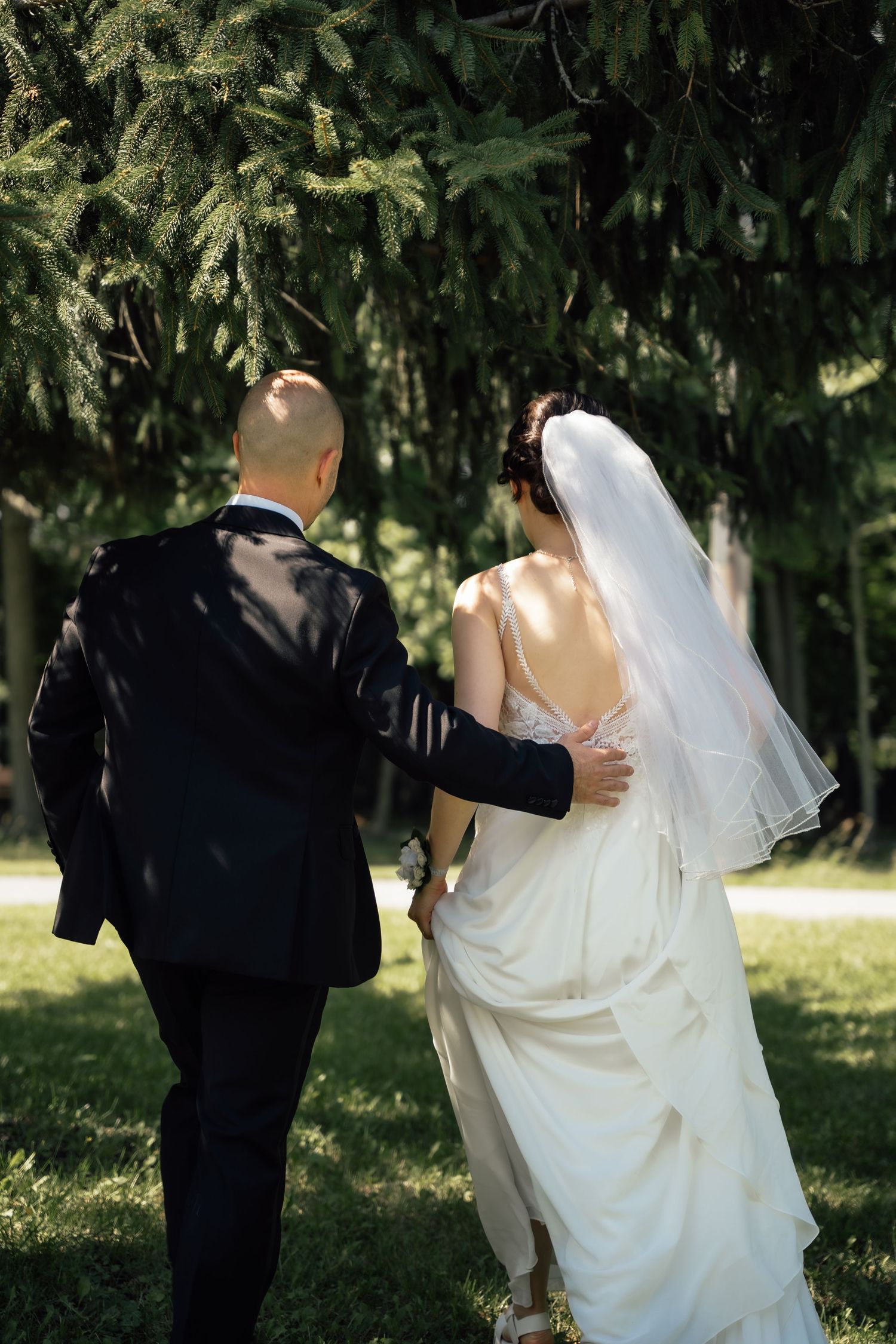Bride and groom walk hand in hand through a sunny green park after their wedding.
