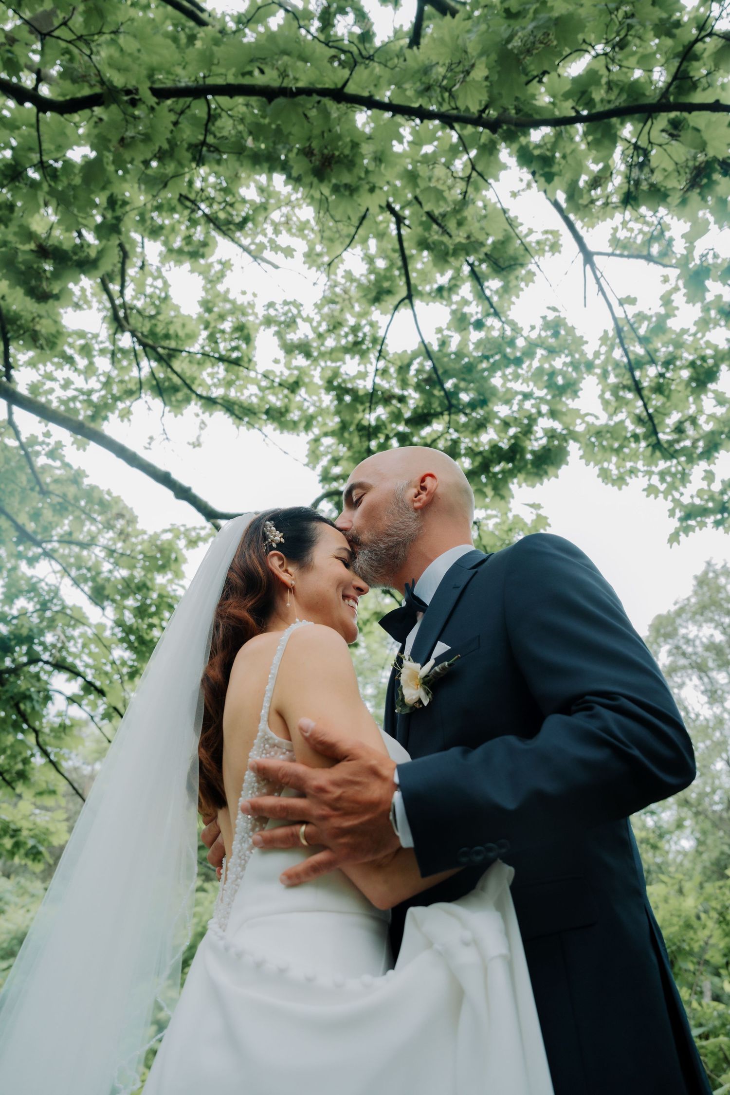 Bride and groom share a romantic moment under lush green trees, with the groom kissing the bride's forehead.