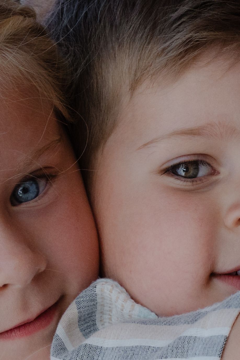 Two young children with blue and hazel eyes pressed cheek to cheek, smiling softly in a close-up portrait photo.
