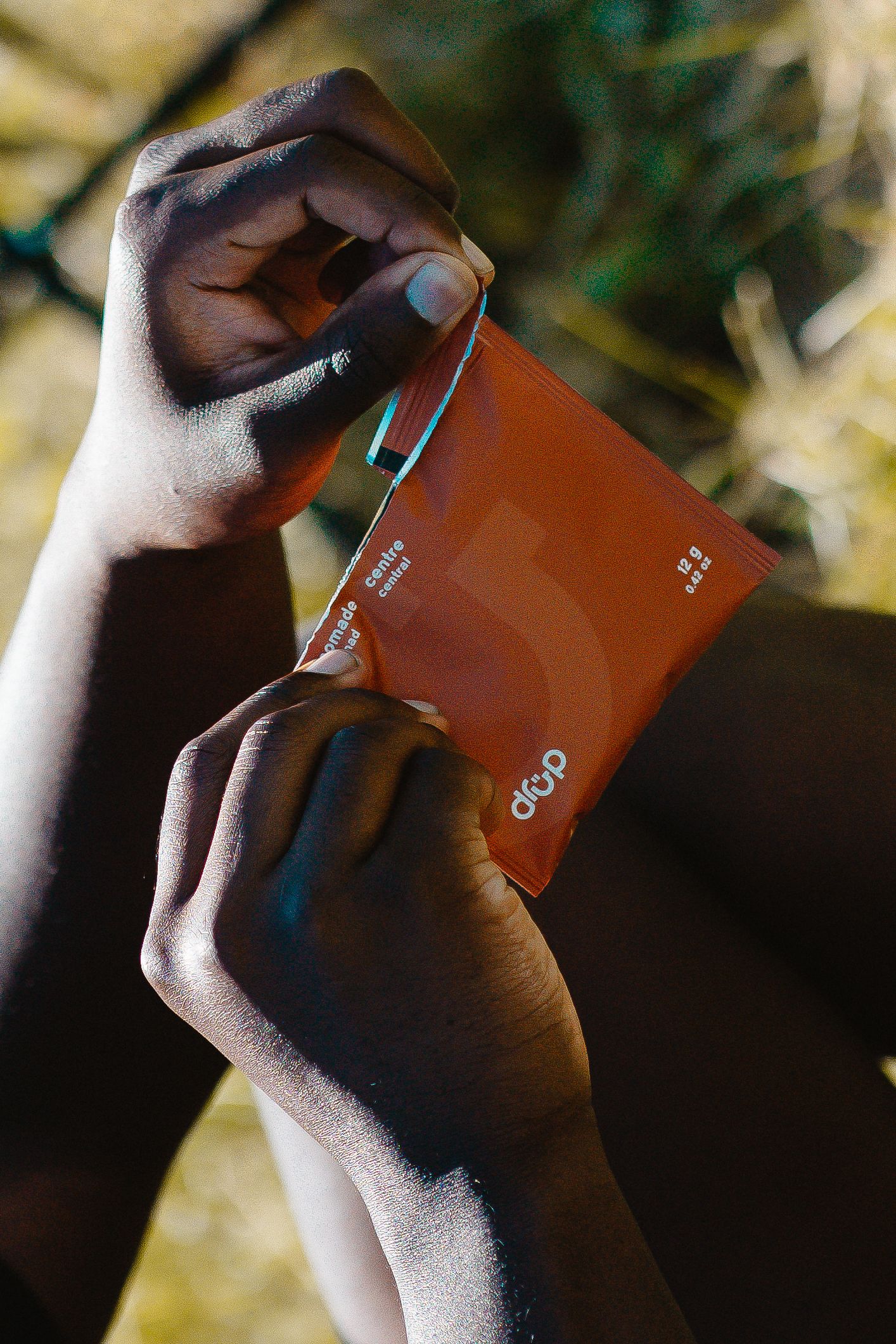 Close-up of dark-skinned hands opening an orange drop brand packet in sunlight outdoors.
