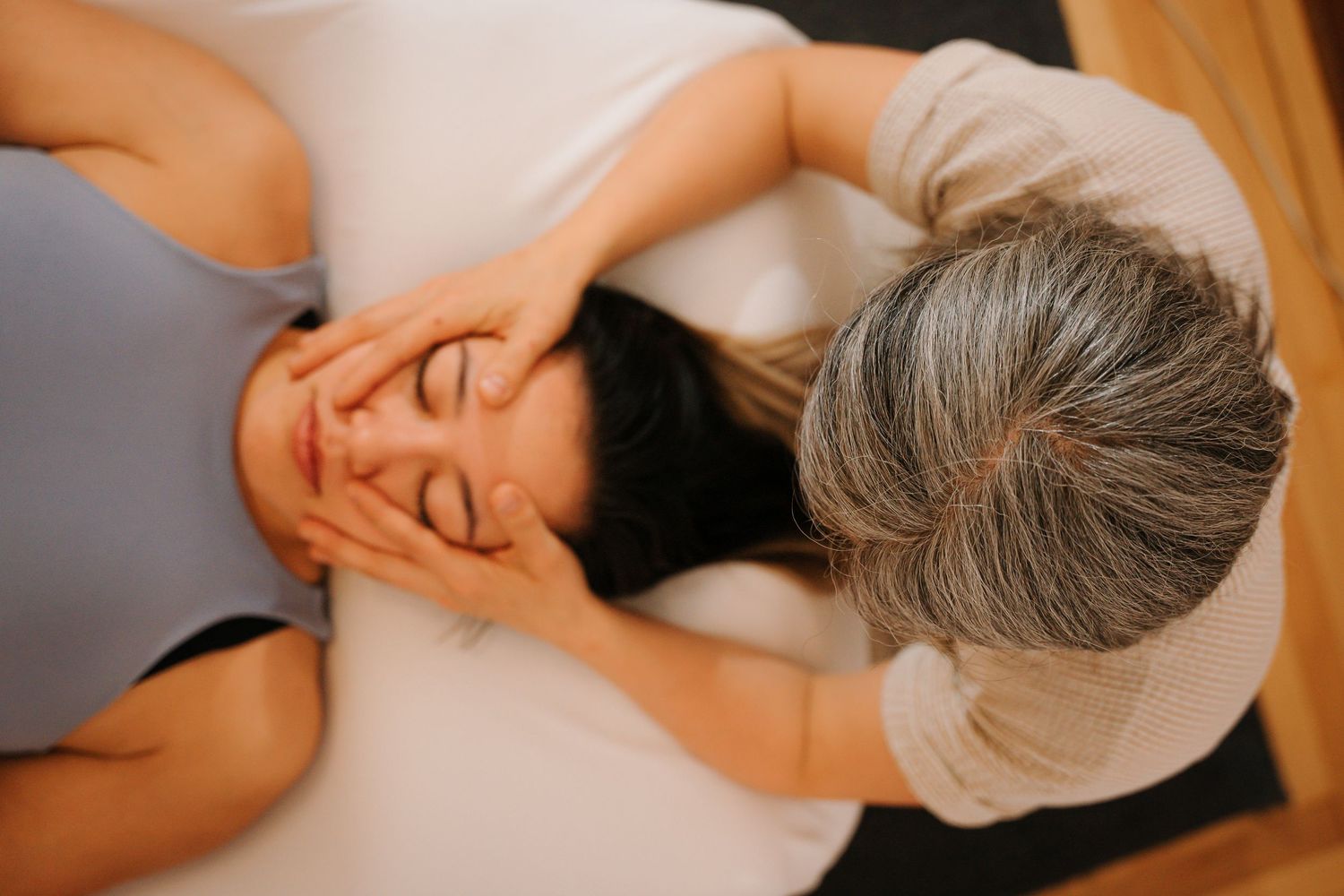 Overhead view of a gray-haired therapist performing a facial massage on a woman lying down.