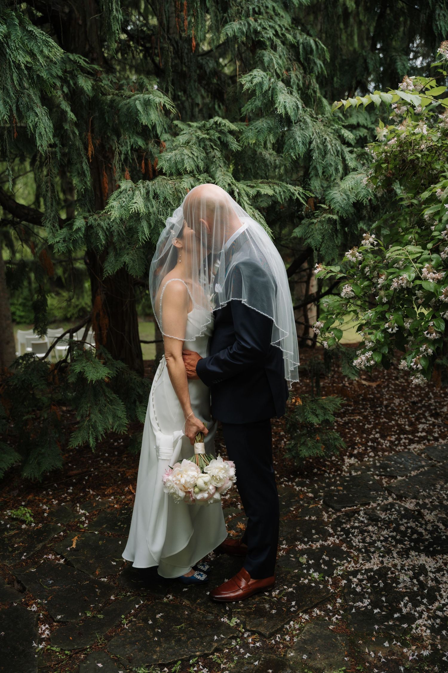 Bride and groom kissing under a white veil in a lush green garden, bride holding white bouquet.