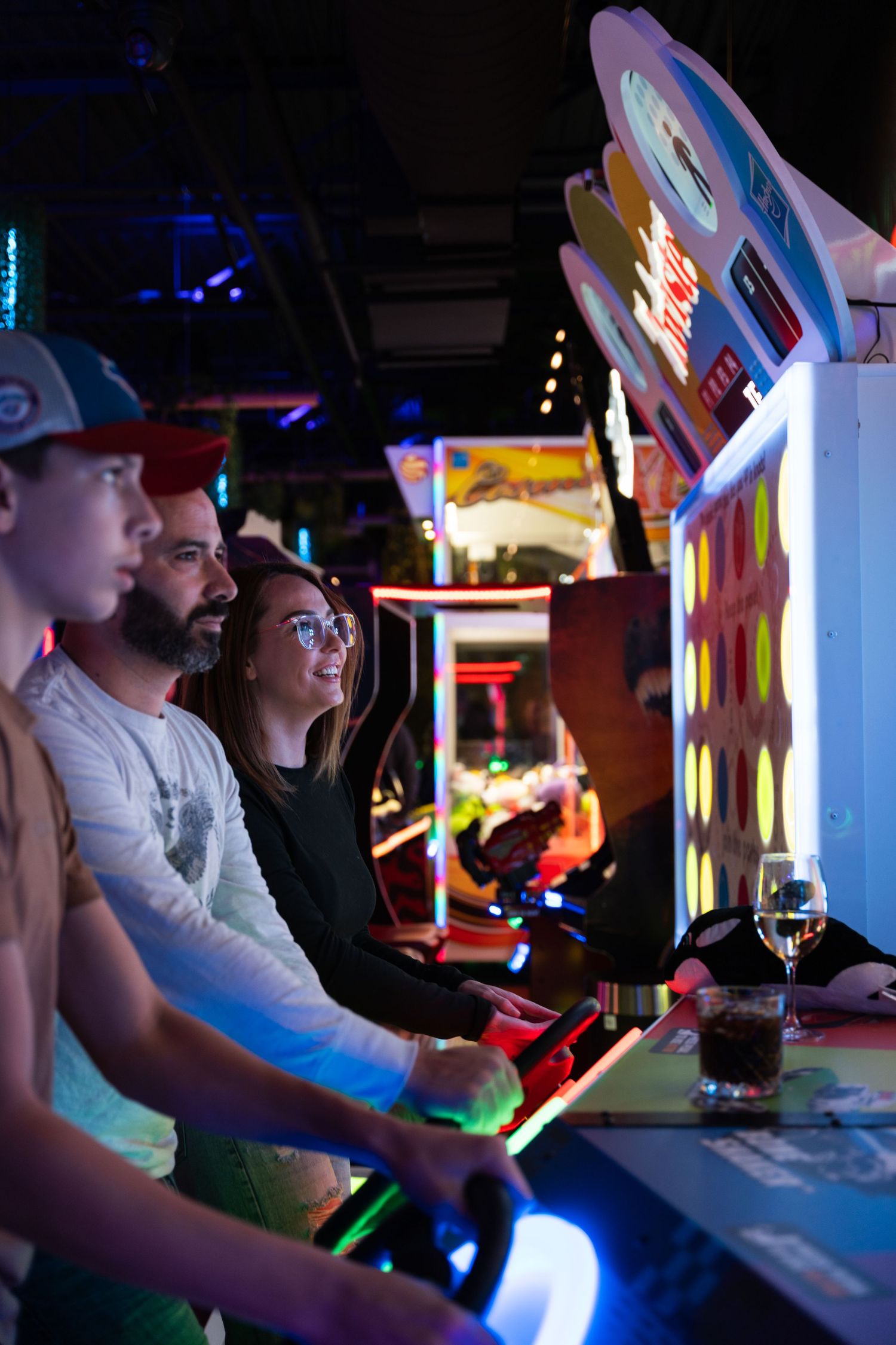 Group of friends playing a racing arcade game with drinks on the console in a dark arcade.