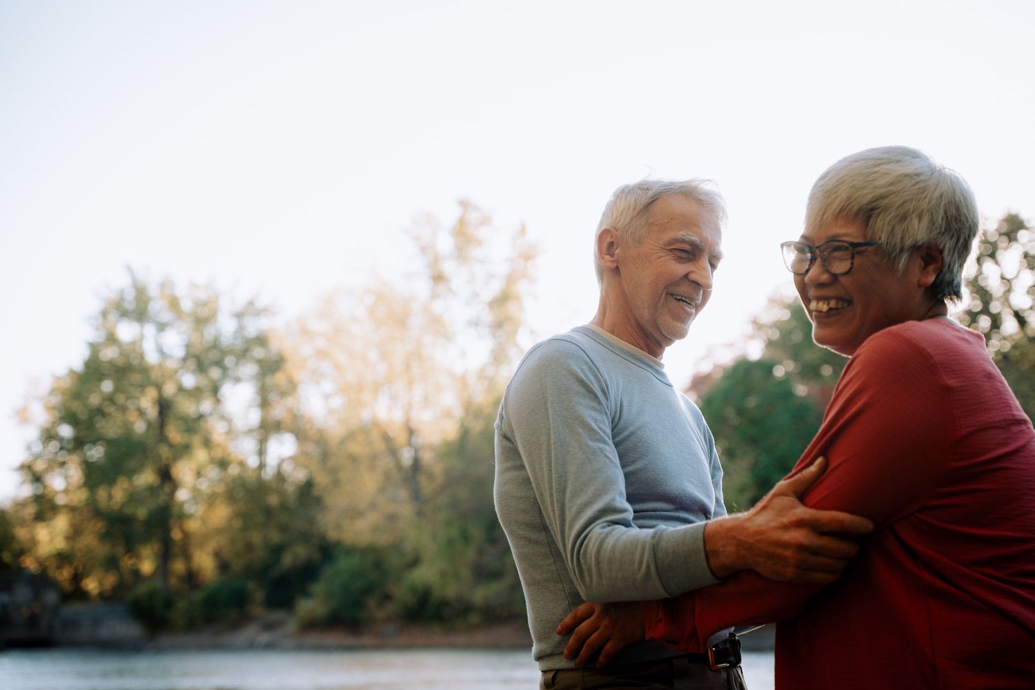 Cheerful senior couple laughing and embracing near a river with colorful autumn trees in the background.
