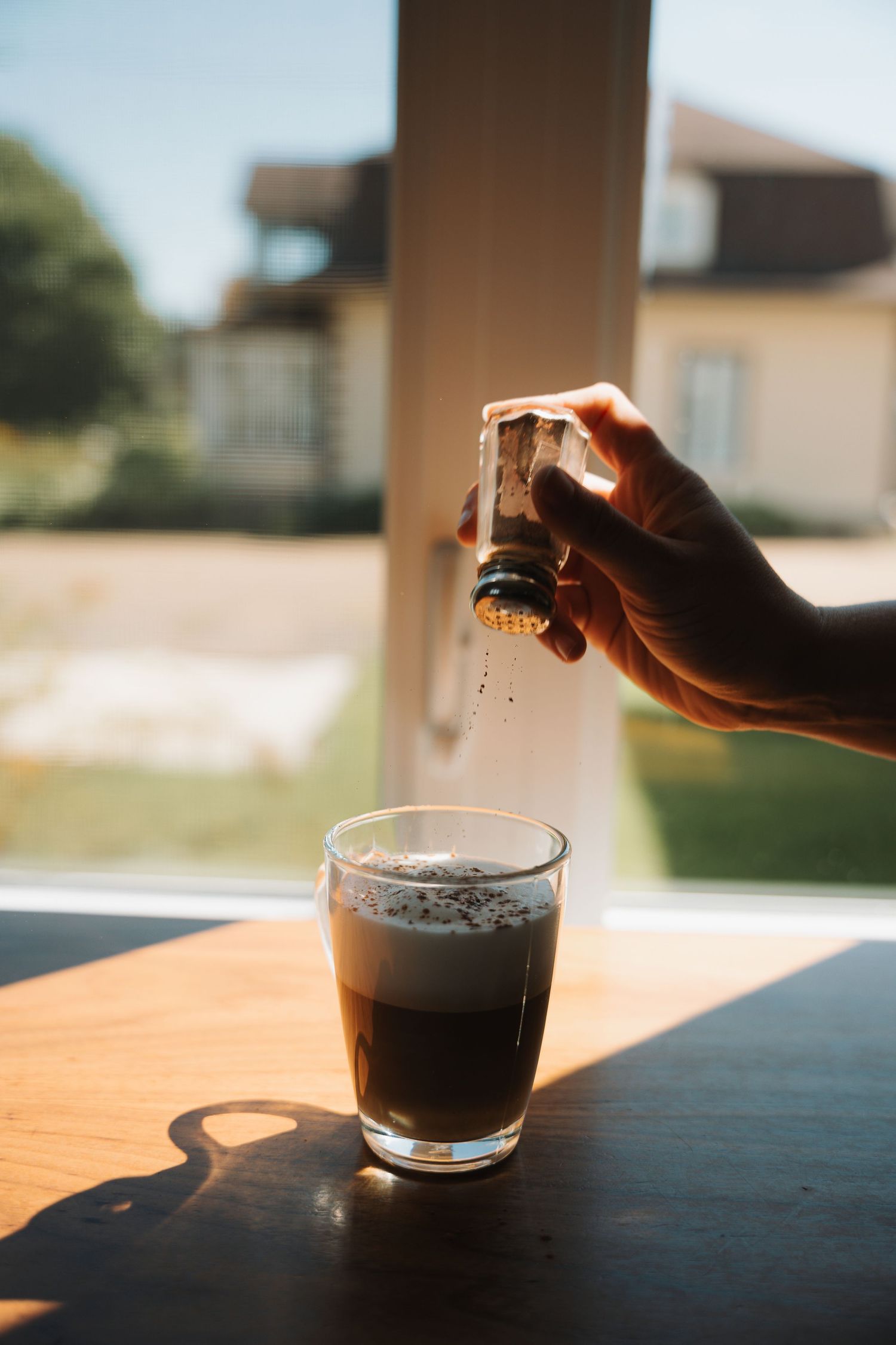 Hand sprinkling spice over a dark layered coffee drink by a sunny window.