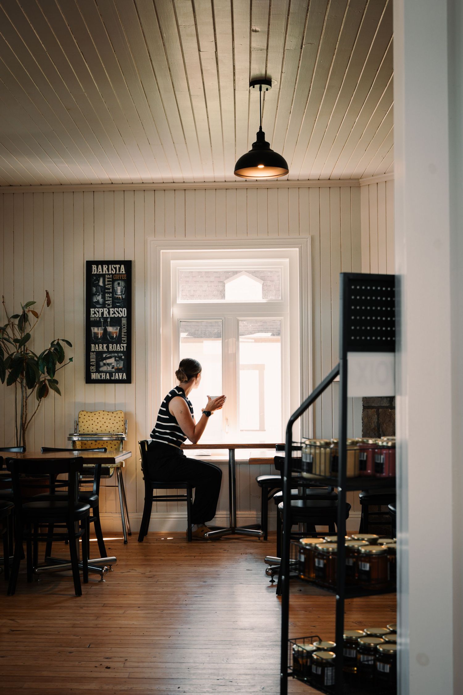 Woman sitting alone by a window enjoying coffee inside a cozy white-walled cafe.