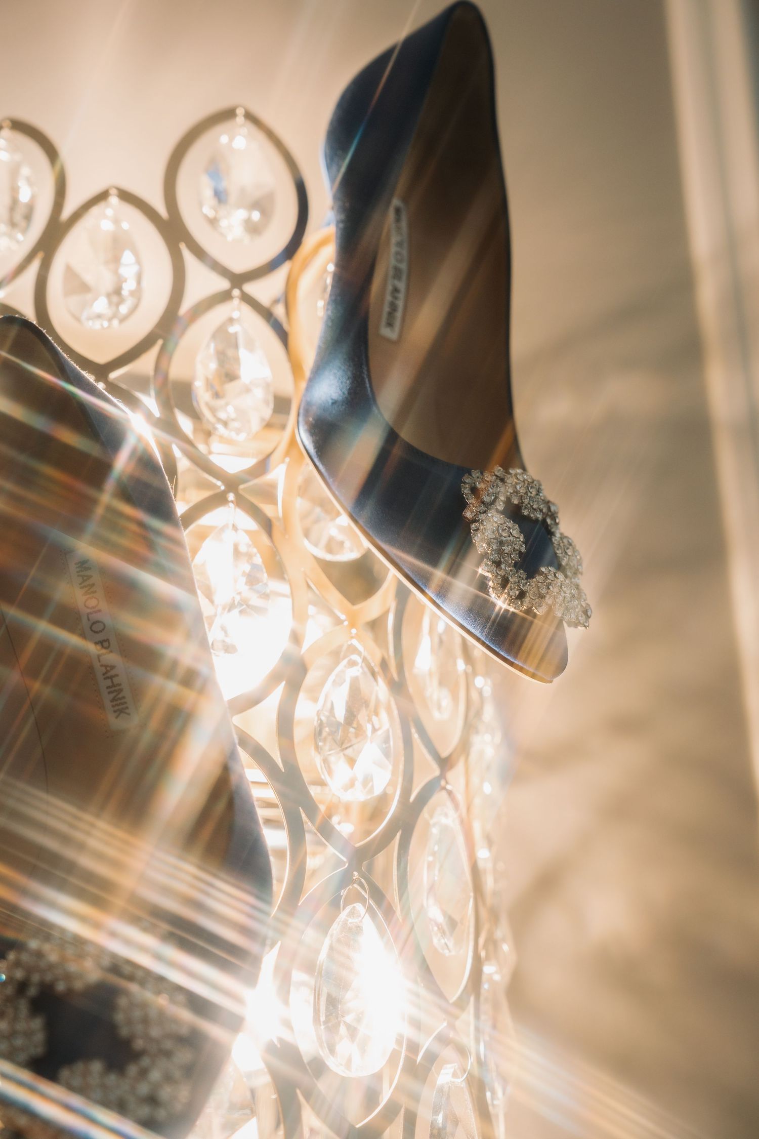Elegant Manolo Blahnik blue satin heels with crystal brooch detail, photographed among sparkling chandelier crystals.