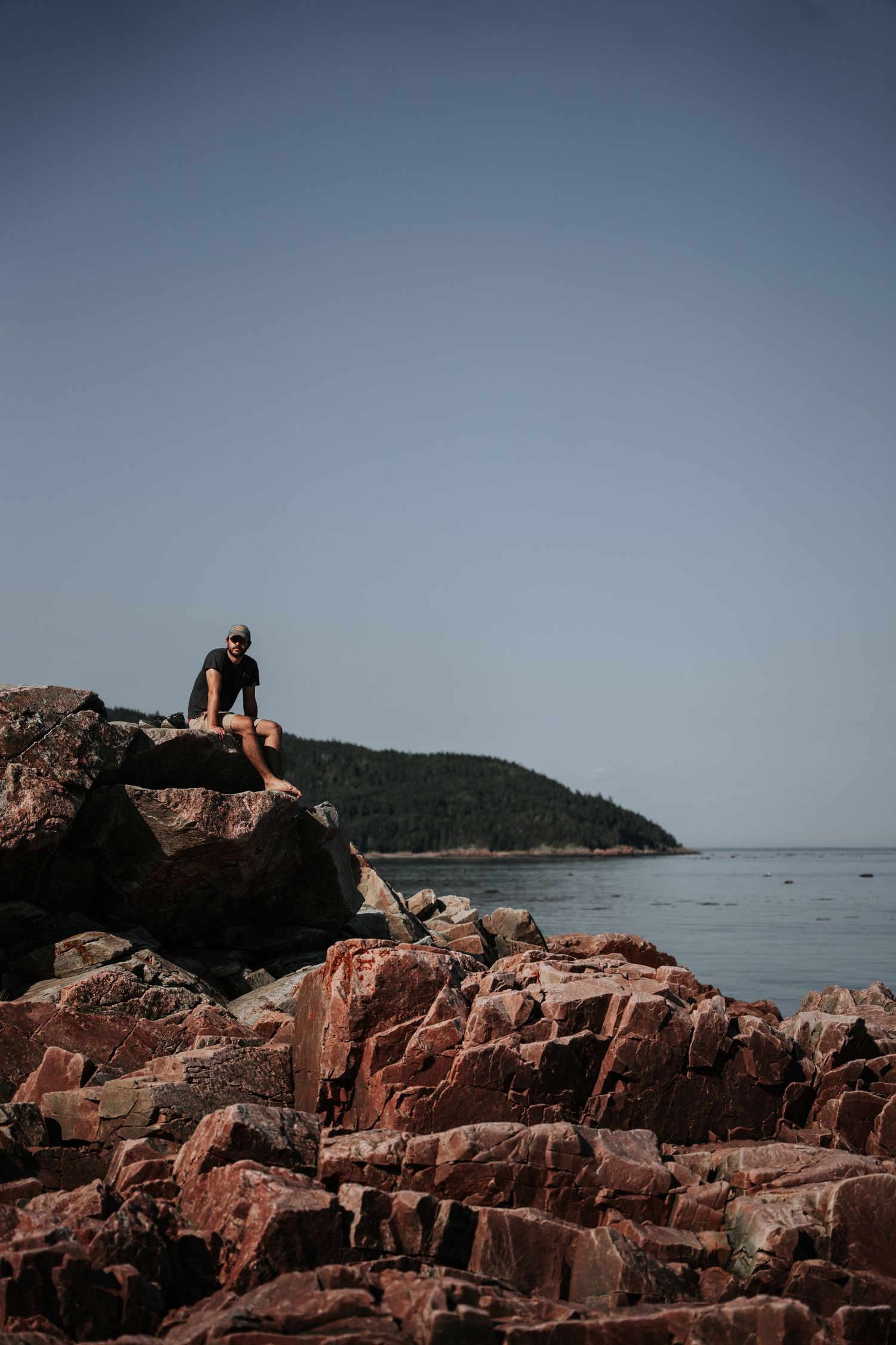 Person sitting on red rocks by the ocean with a forested island in the background on a clear day.