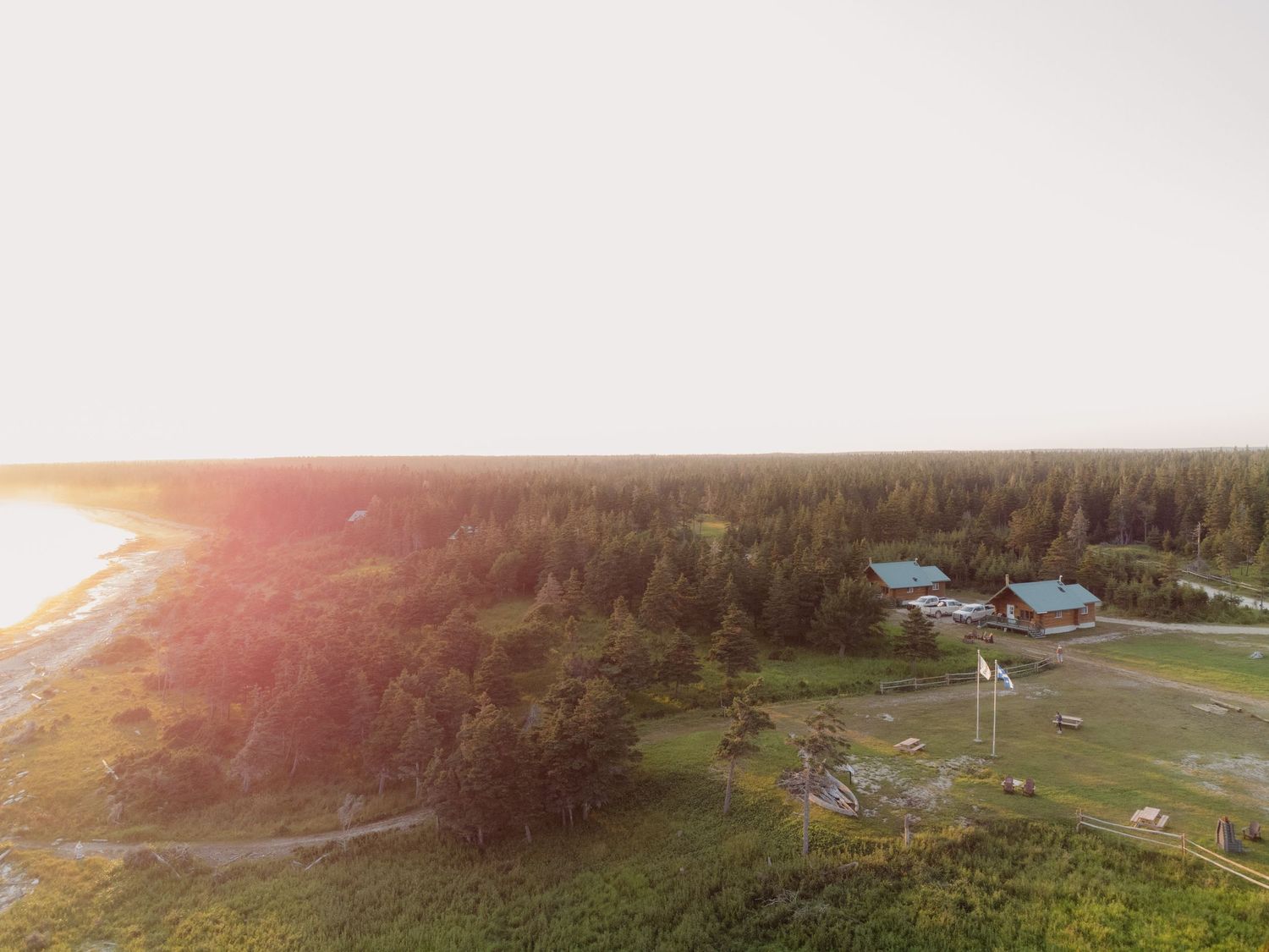 Aerial sunset view of a remote wilderness lodge surrounded by dense boreal forest near a lake shoreline.