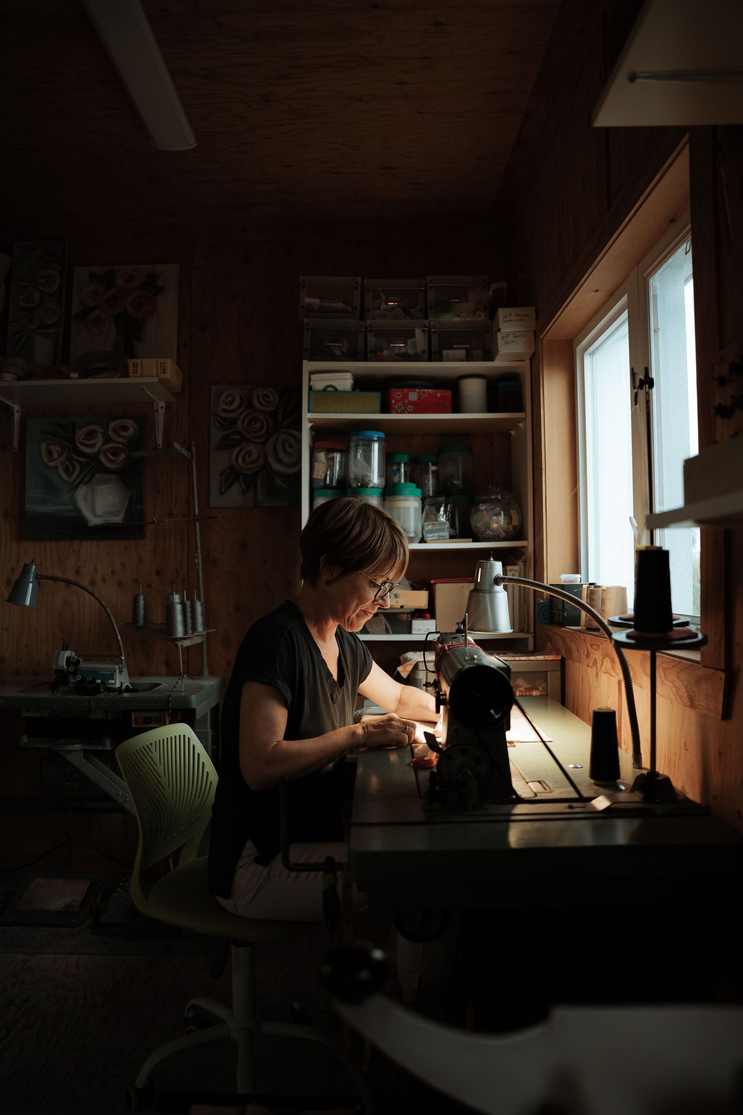 Woman working at a sewing machine in a dimly lit wooden craft studio by a window.