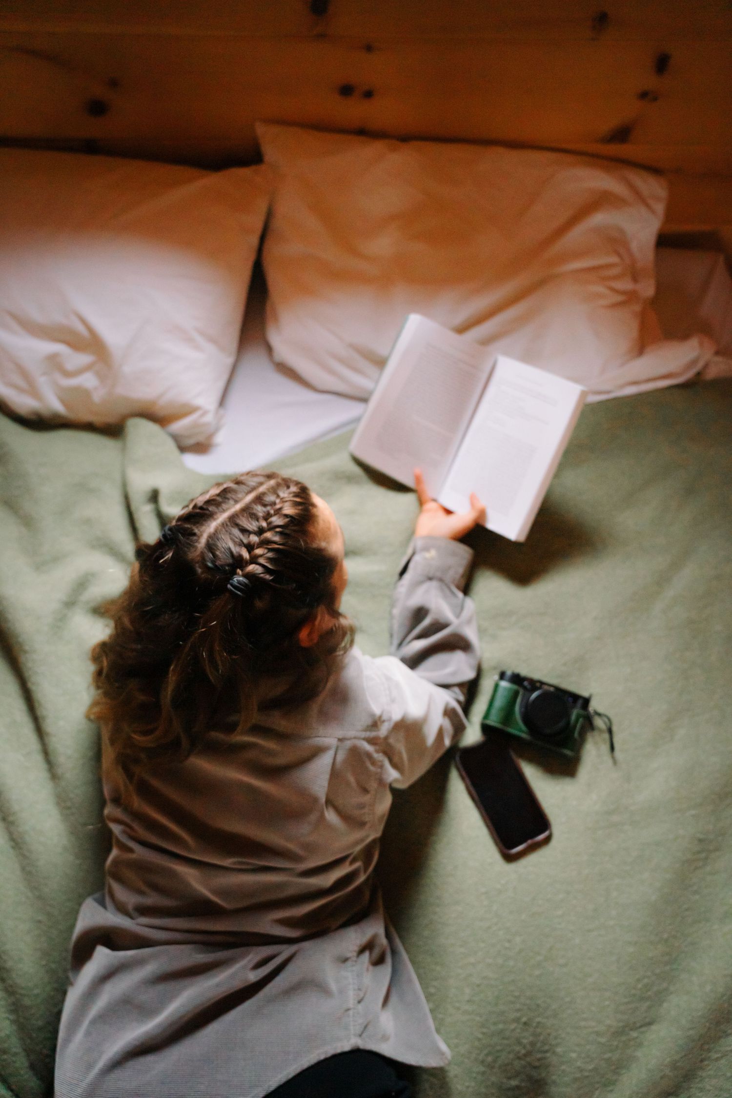 Overhead view of a girl with braided hair reading a book on a green bed with a camera nearby.