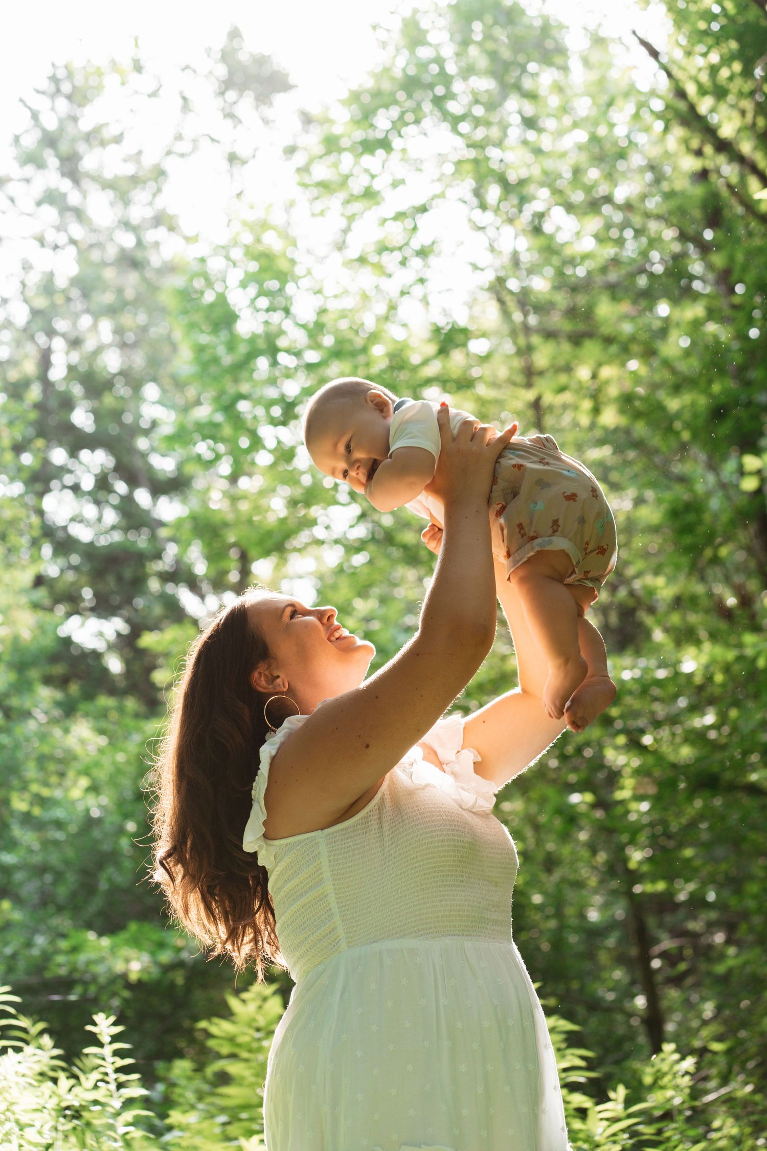 A joyful mother in a white dress lifts her smiling baby overhead in a sunlit green forest.