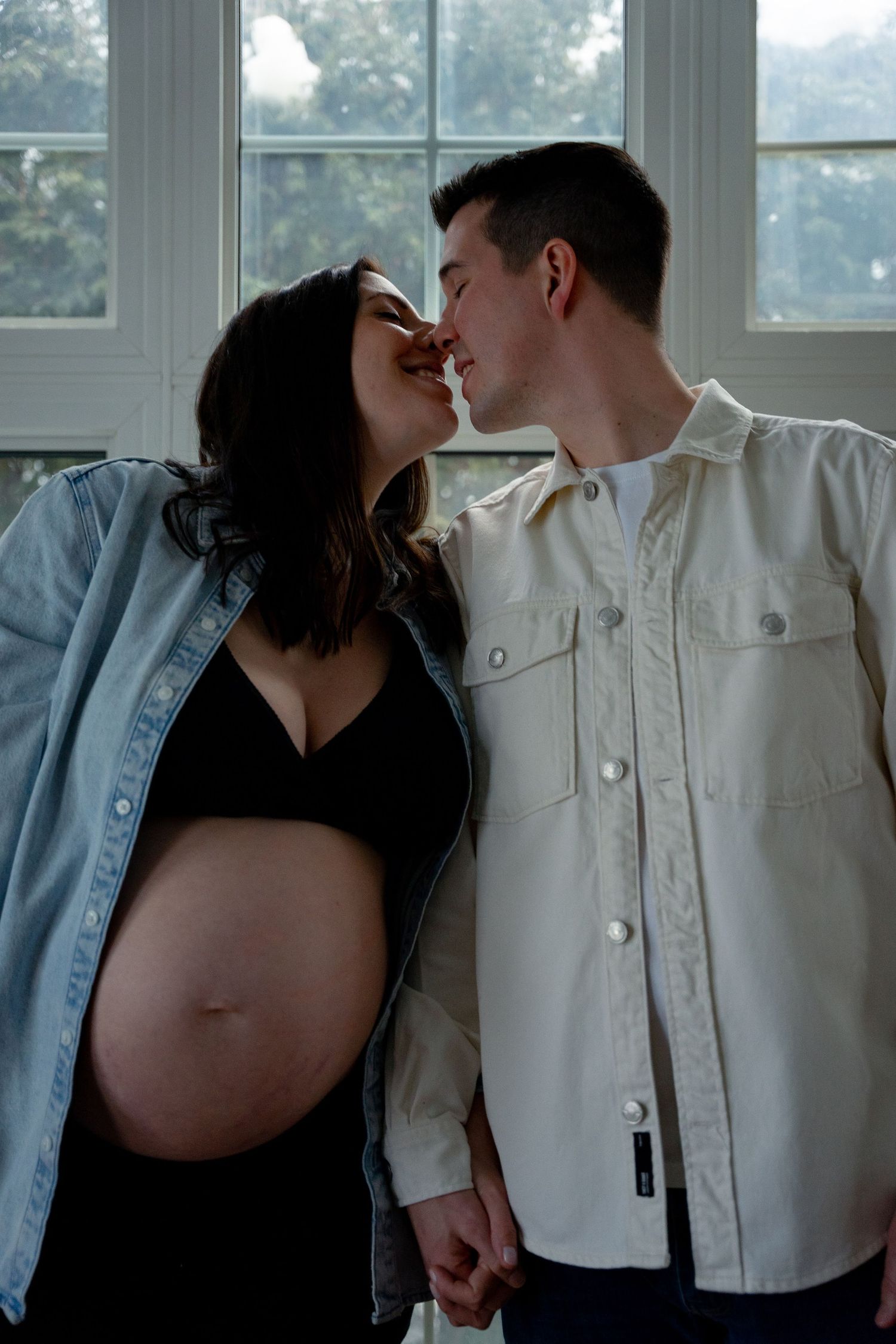 Pregnant couple holding hands and nearly kissing in front of a large bright window.