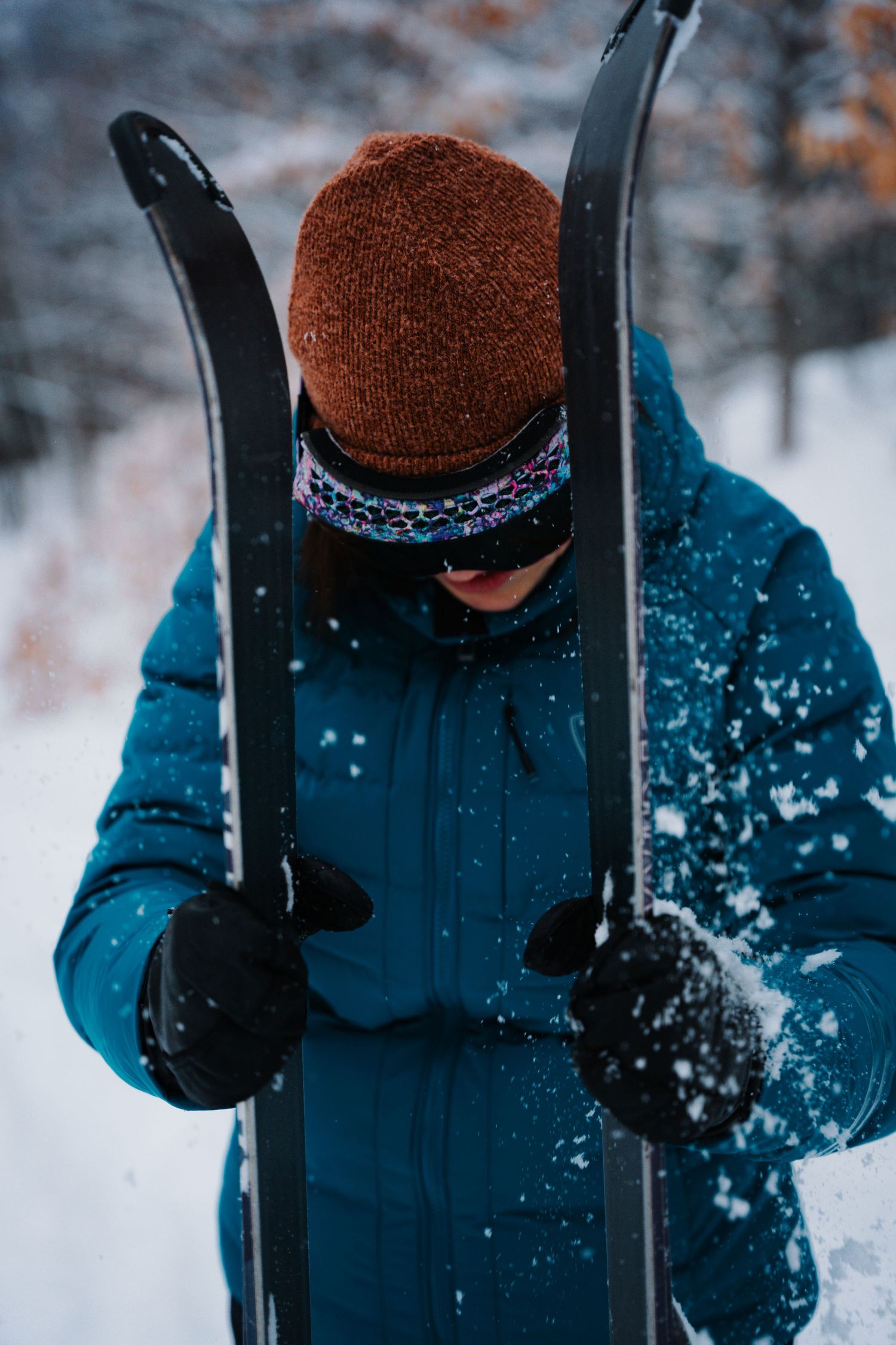 Skier in blue jacket and brown beanie holding skis in snowy winter forest setting.
