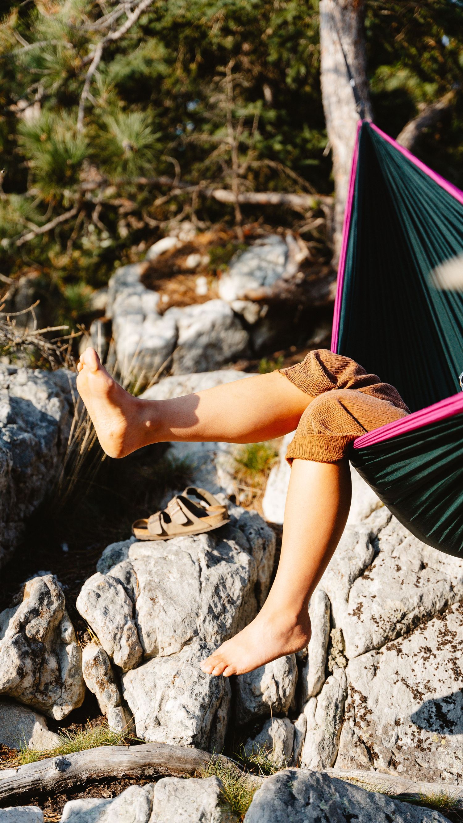 Person relaxing in a colorful hammock with legs dangling over rocky terrain in a forest setting.