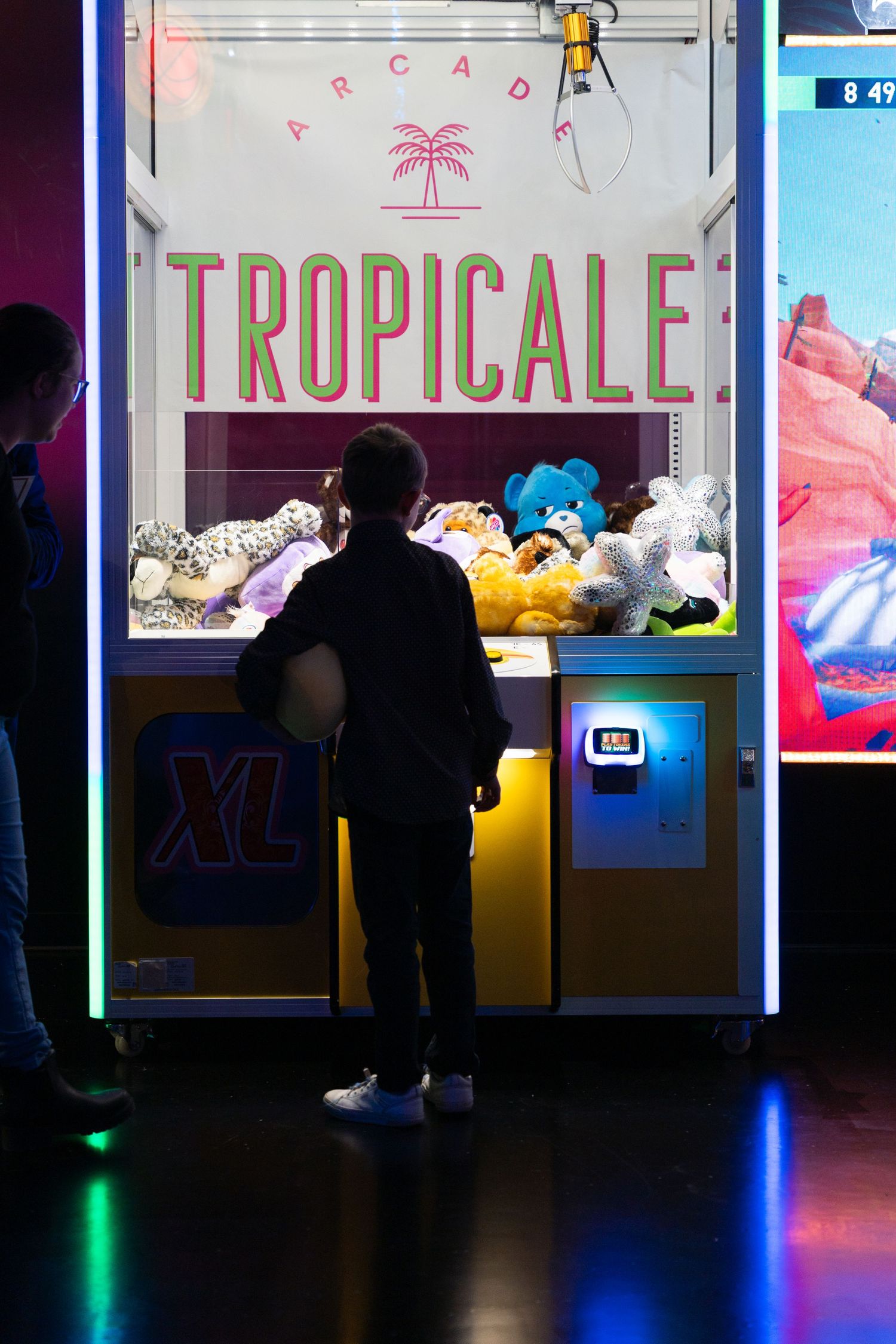Child stands before a large Arcade Tropicale claw machine filled with stuffed animals.
