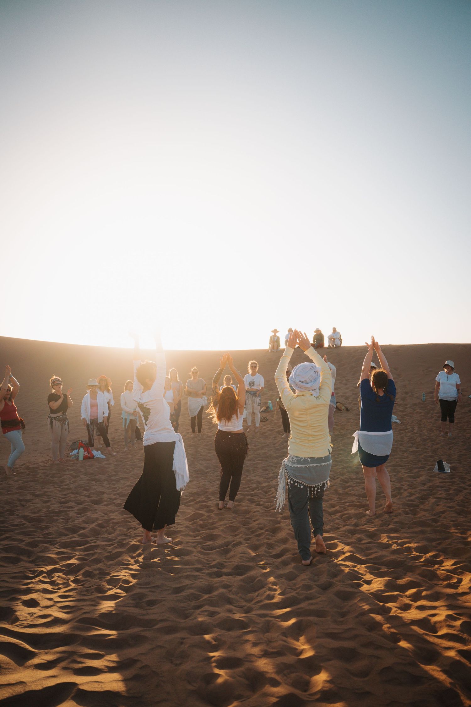Large group of people raising arms toward the sunset during an outdoor desert gathering.