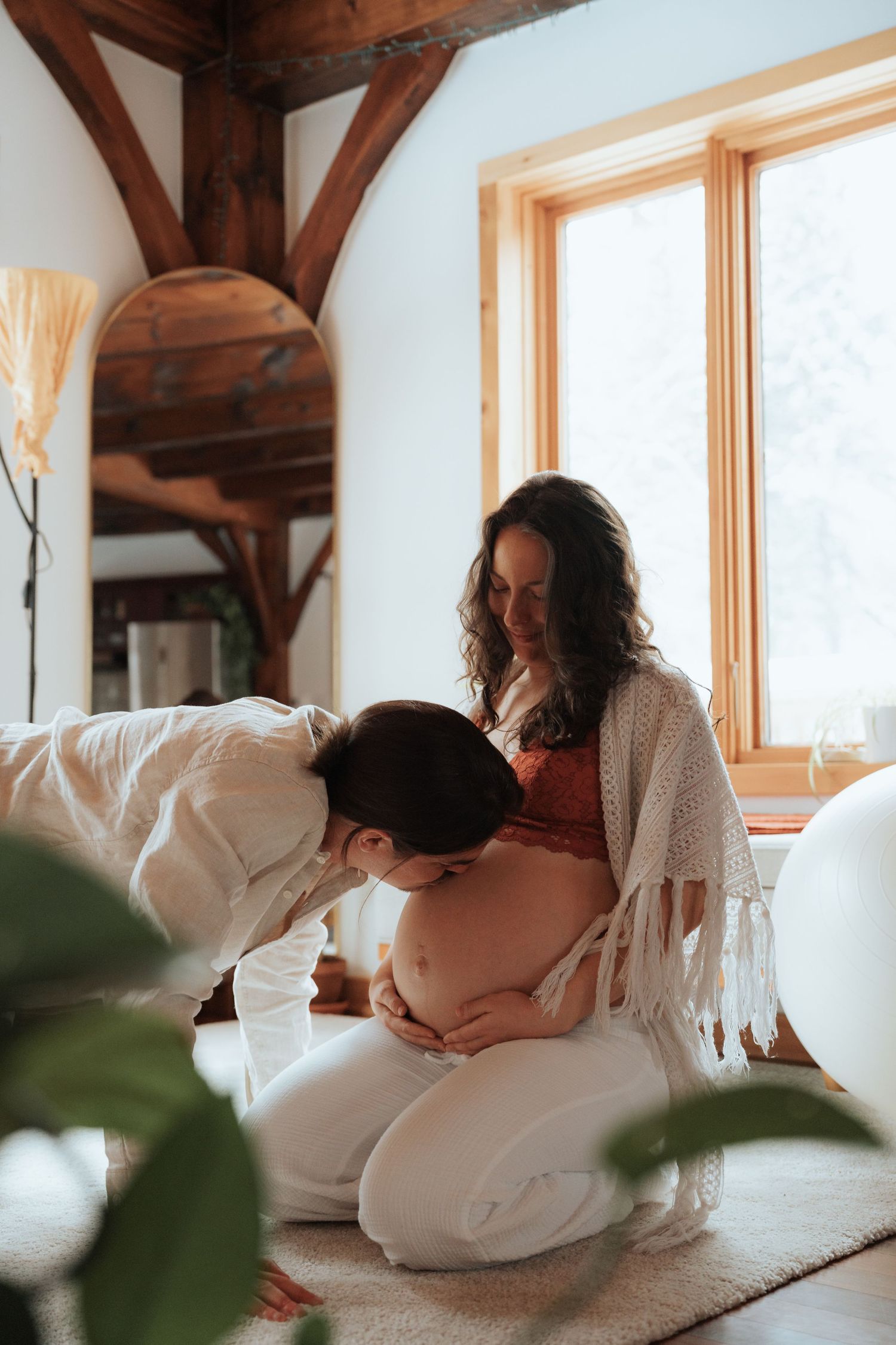 Partner kissing pregnant woman belly while she kneels on floor in bright wooden cabin interior.