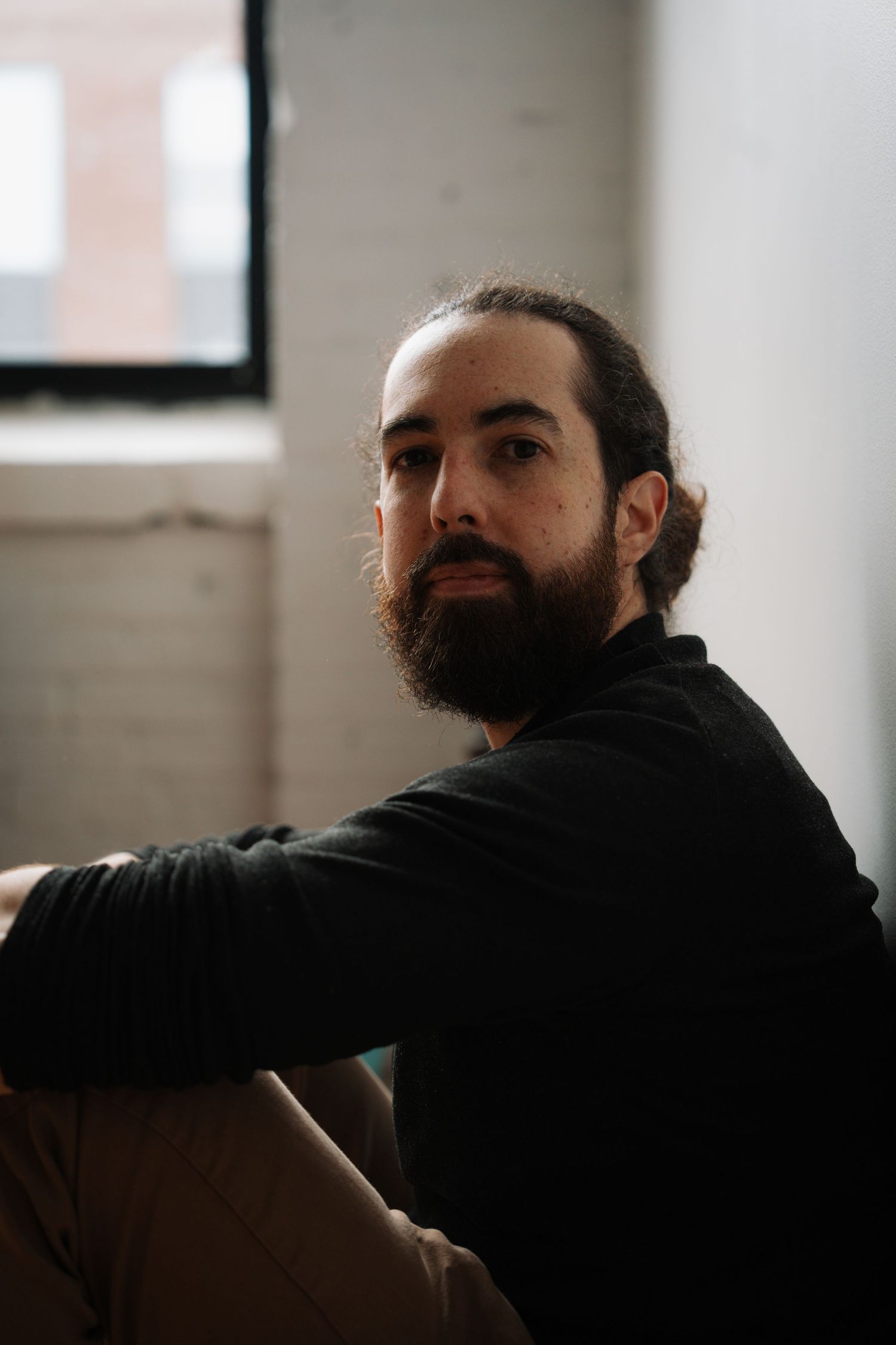 Bearded man with long hair in a bun wearing a black turtleneck seated casually indoors.